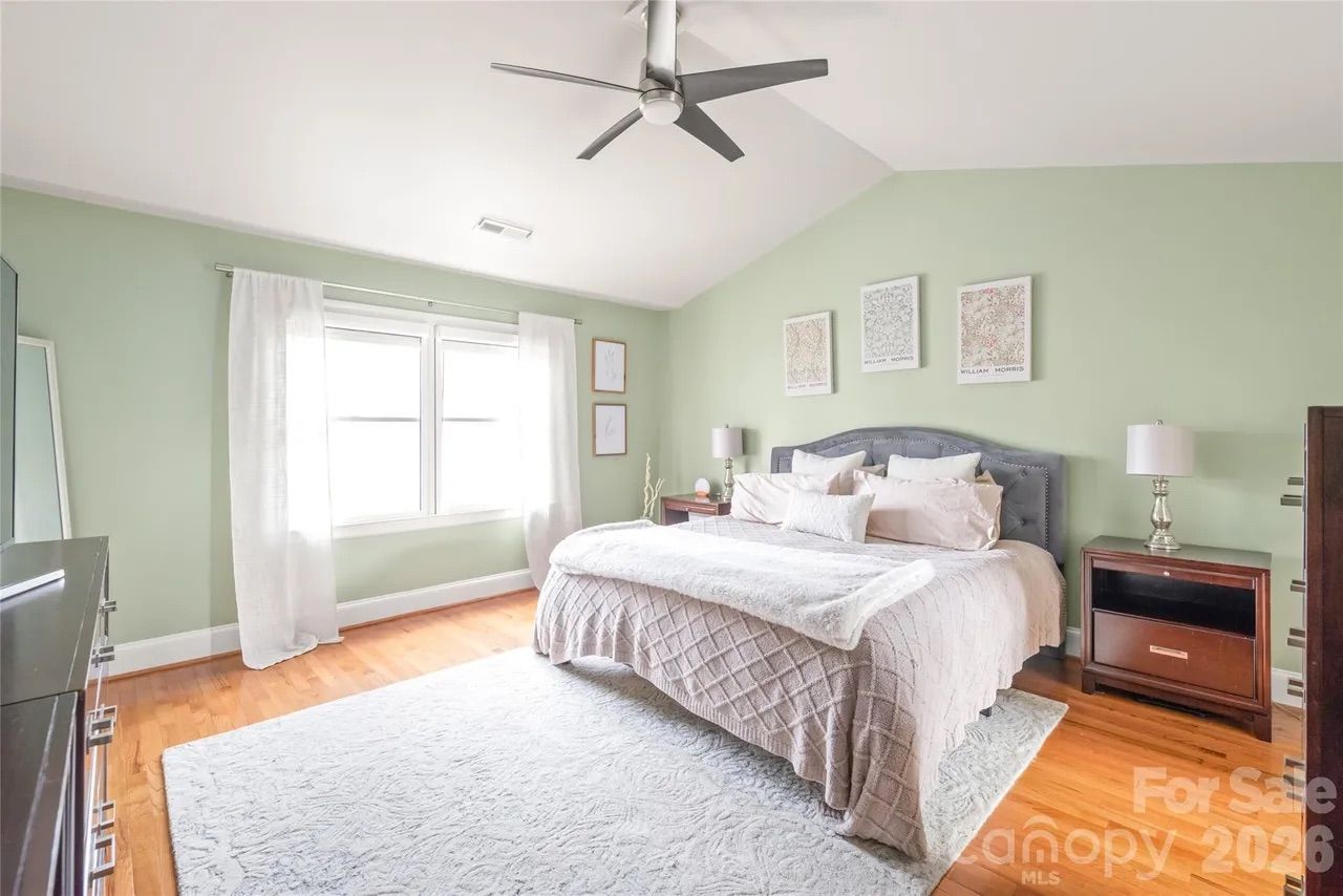Bright bedroom with light green walls, large window with white curtains, king bed with gray headboard and beige bedding, wooden floor, white rug, ceiling fan, and dark wood nightstands with lamps.