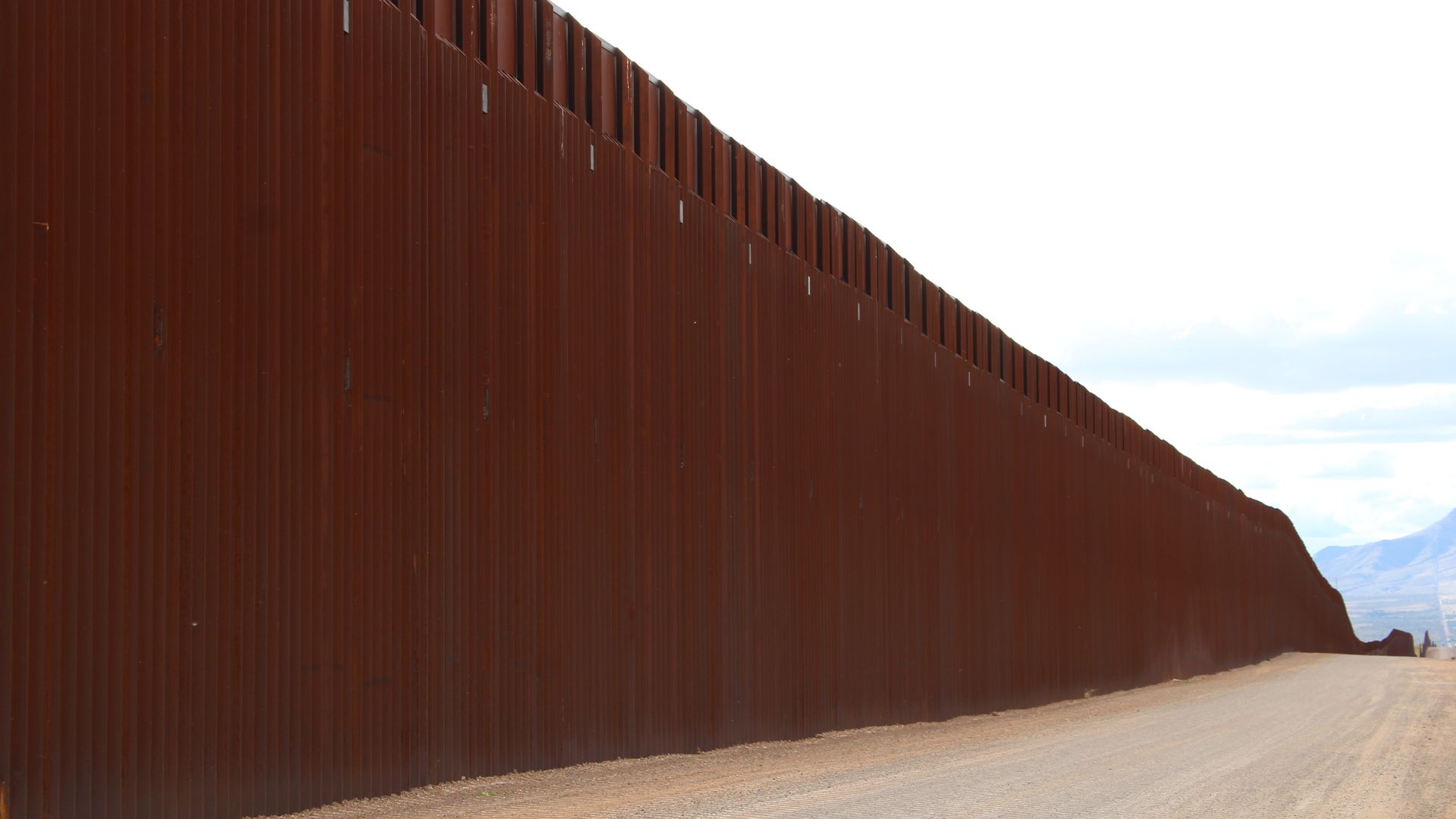Tall rust-colored metal border wall extending along a dirt road under a bright, partly cloudy sky with mountains in the distance.