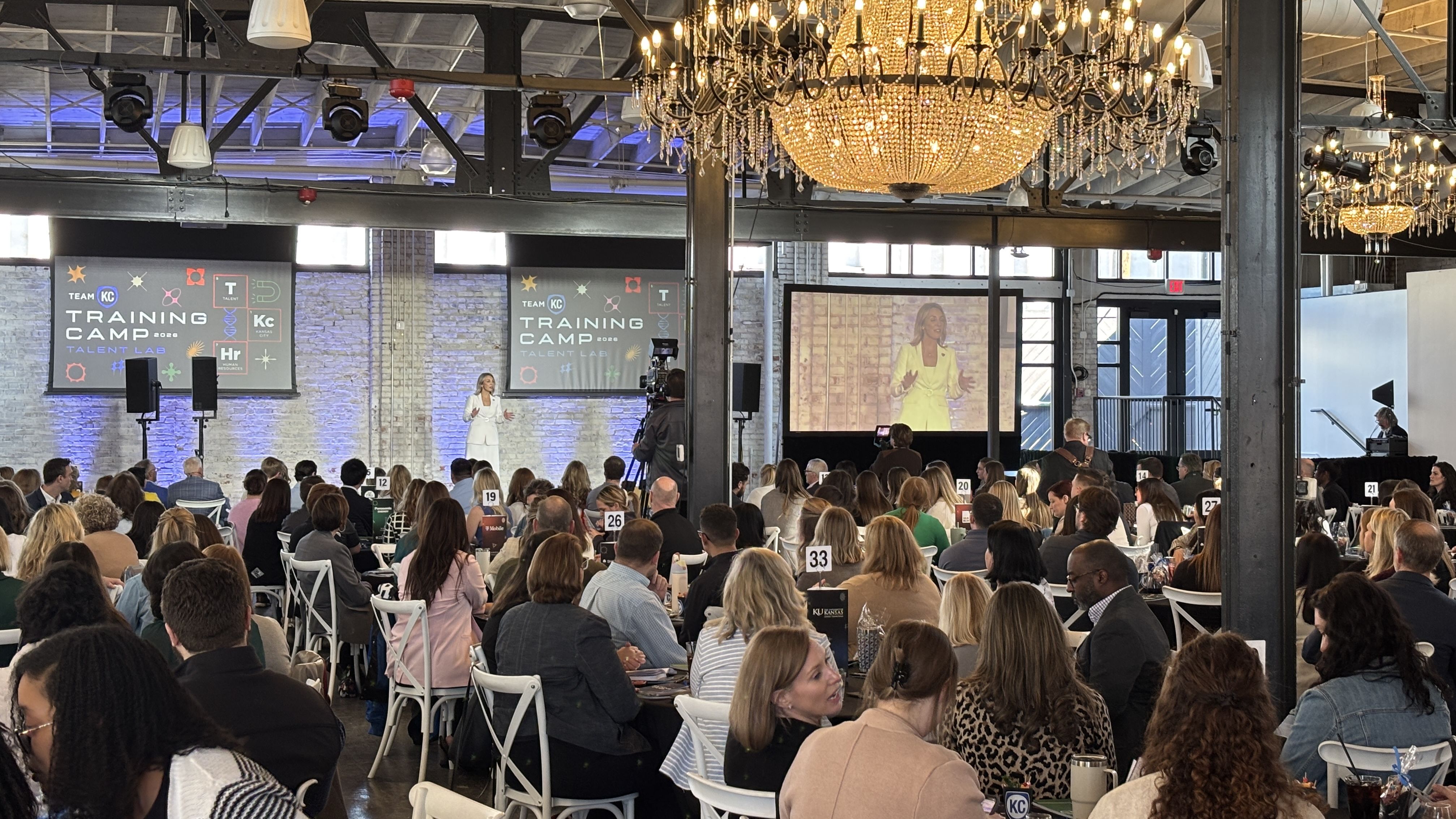 Crowd of people seated at round tables in a large room with chandeliers, watching a woman in white speaking on stage at the Team KC Training Camp 2026 Talent Lab event.