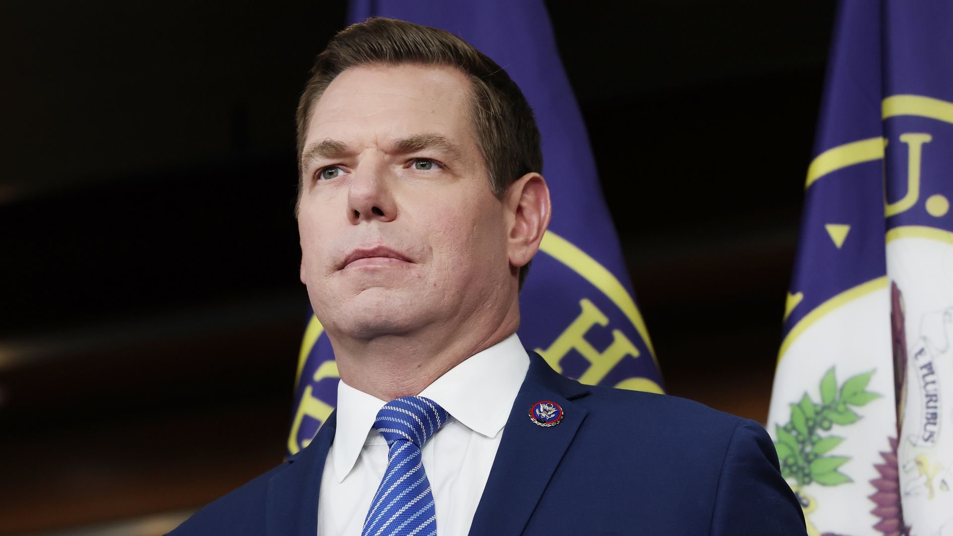 A dark-haired Rep. Eric Swalwell,  wearing a navy jacket with a pin at the top of his left lapel, white shirt and blue tie with yellow stripes, stares ahead in front of two flags.
