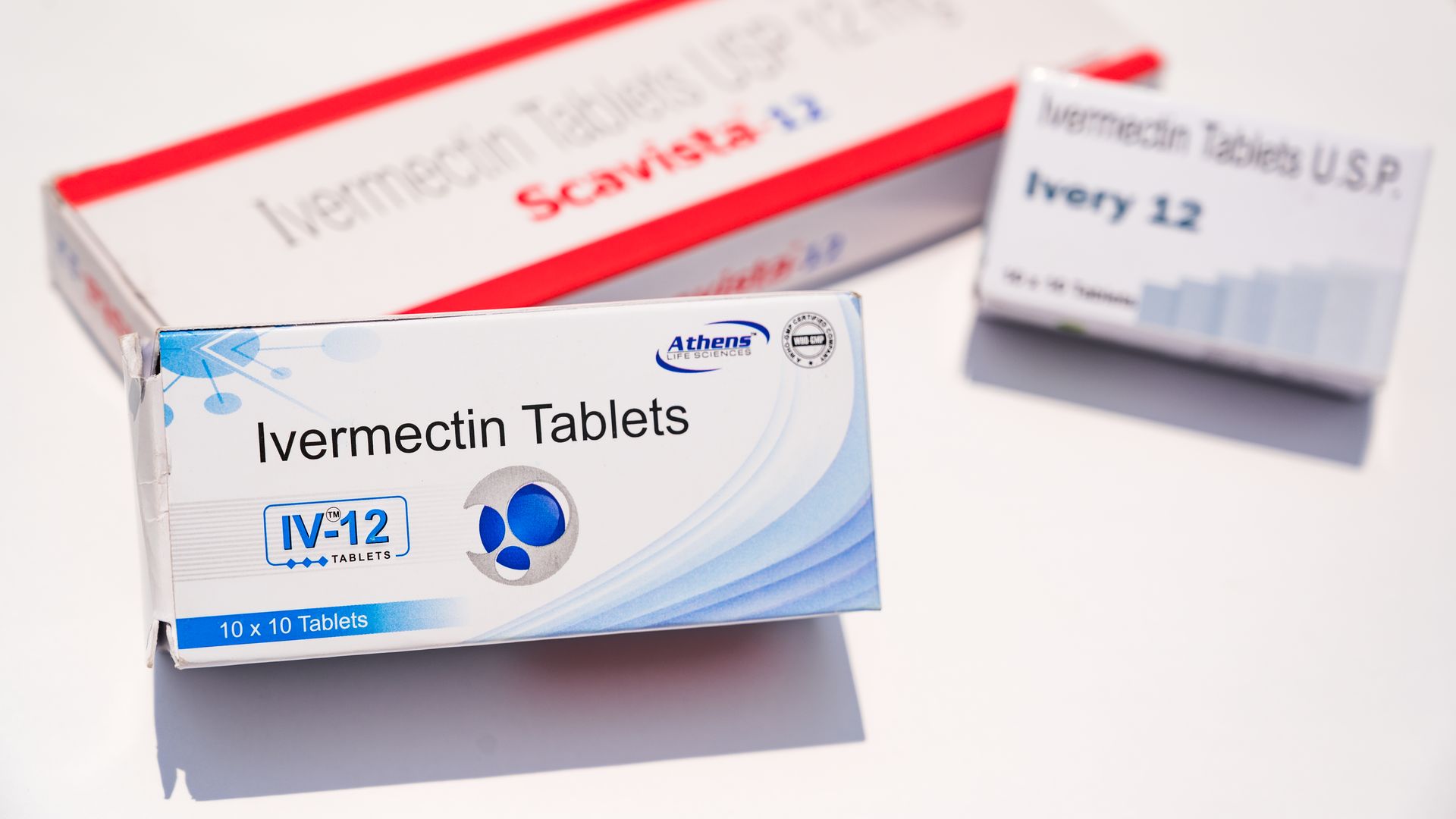 Three boxes of Ivermectin tablets on a white surface, with packaging in white and blue, and red and white, labeled with different brand names and dosages.