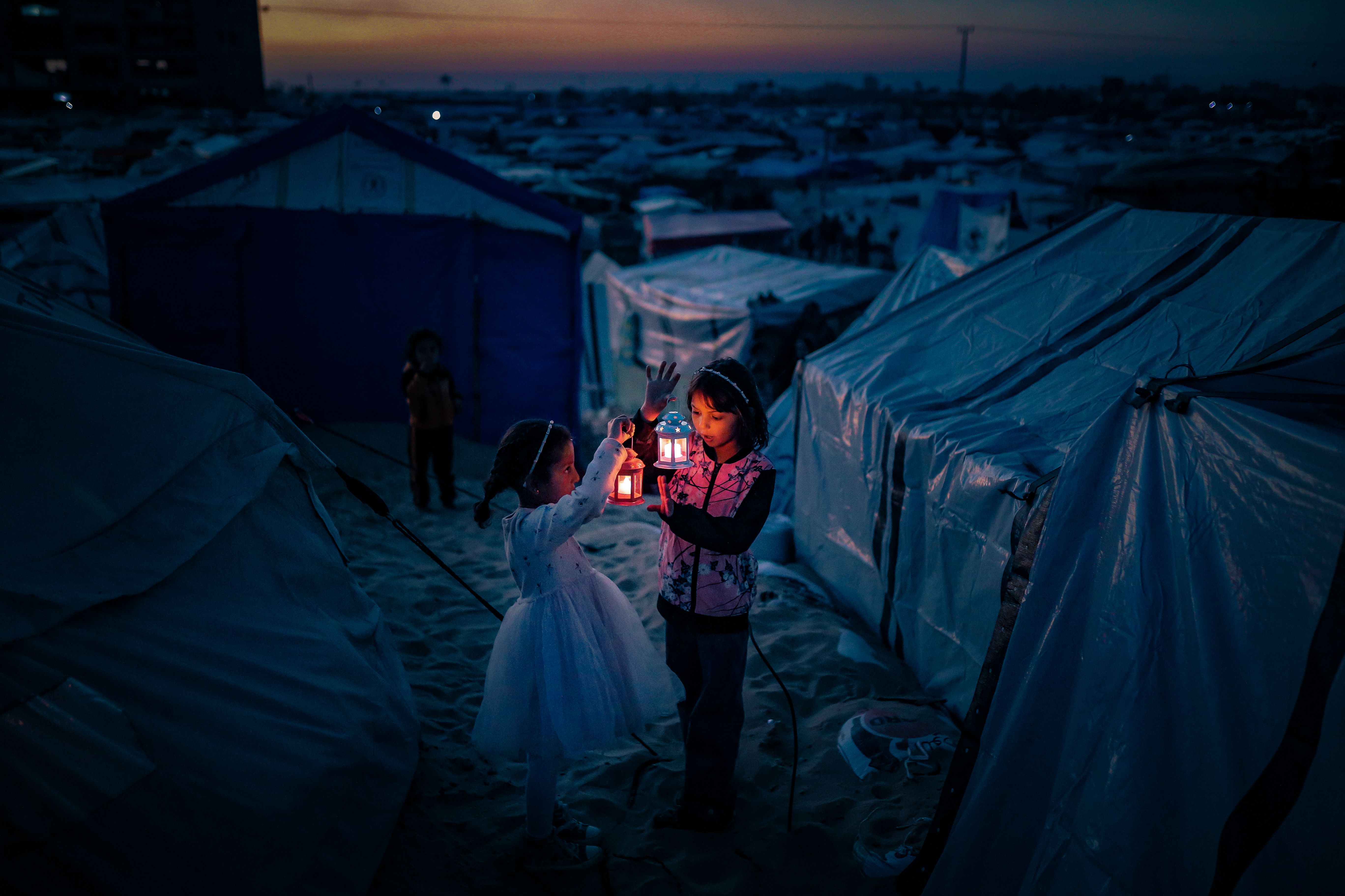Two Palestinian children in the evening, standing between refugee tents and hold glowing Ramadan lanterns. 