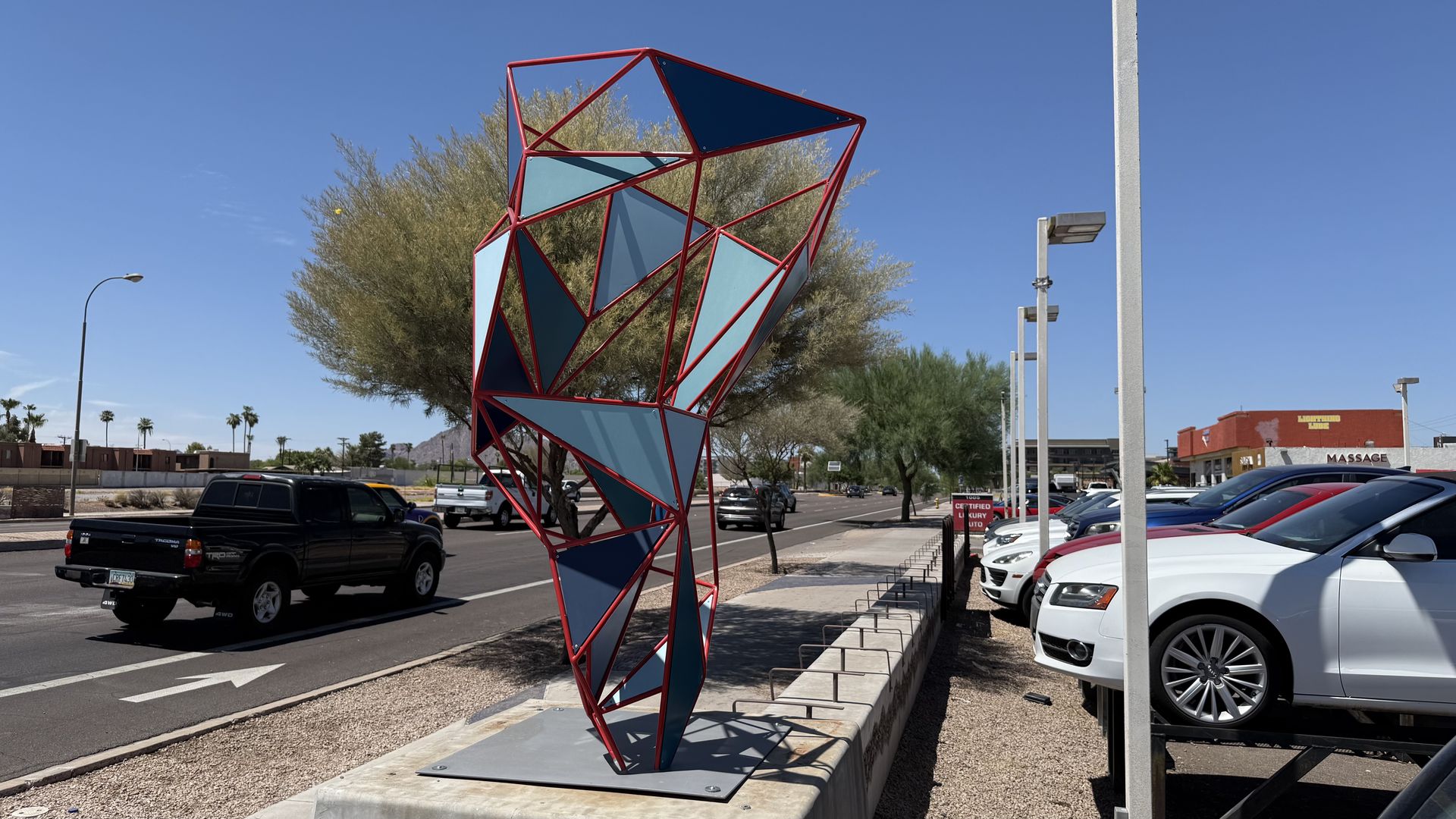 Red metal sculpture with blue and dark blue triangular panels stands on a concrete base beside a street.