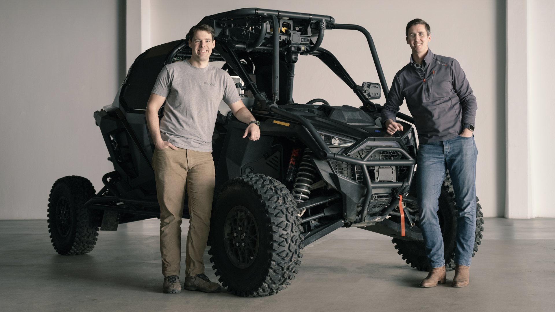 Two men stand beside a large, autonomous ground vehicle. They are dressed casually. The photo looks like it was taken in a showroom or a photo studio.