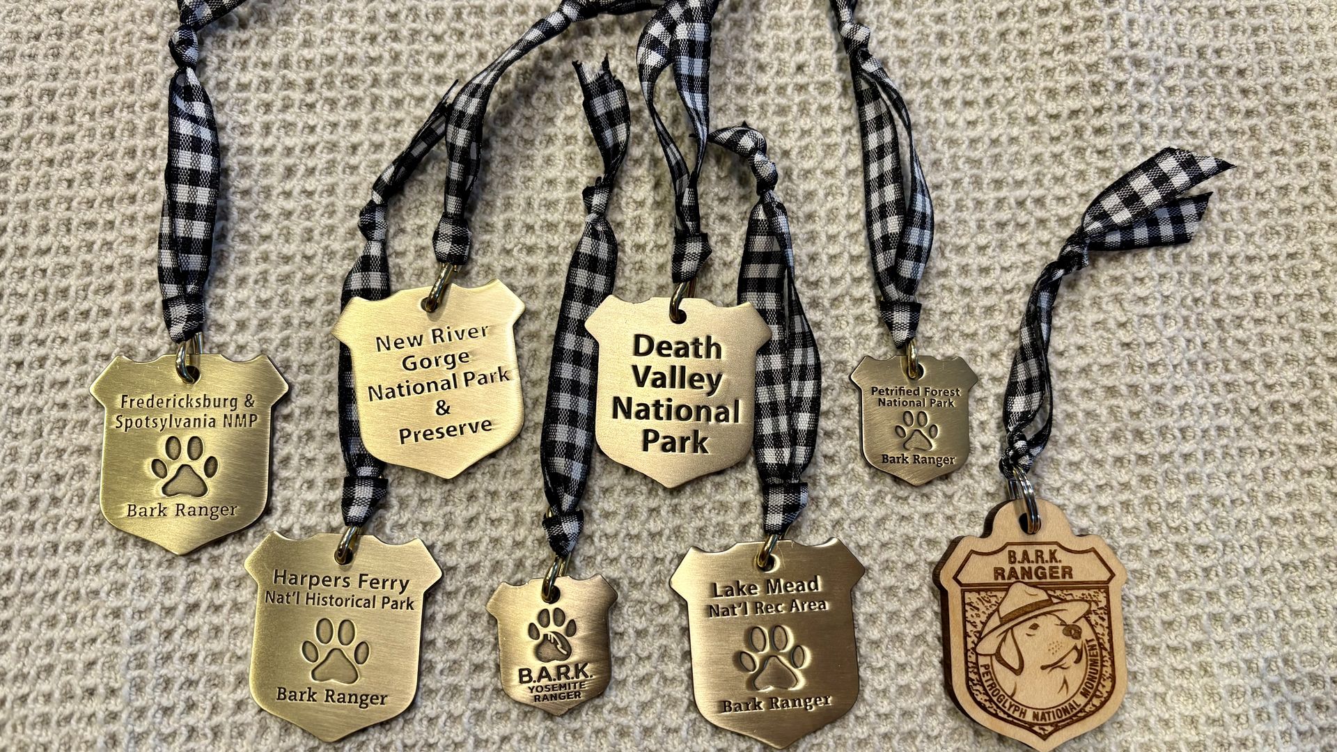 Eight gold and one wood shield-shaped Bark Ranger tags with black-and-white checkered ribbons, each engraved with names of U.S. national parks and preserves, displayed on a textured fabric.