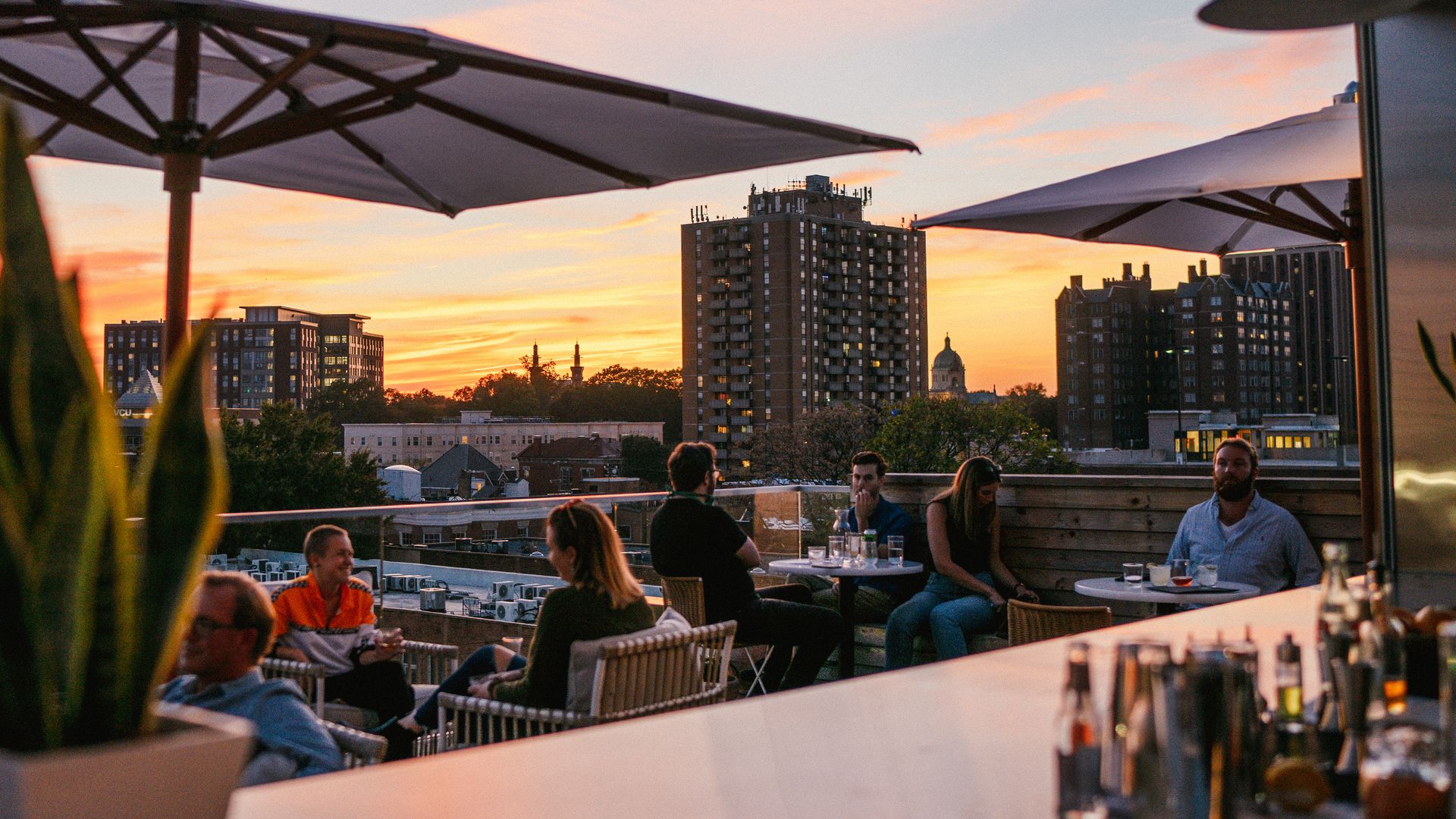 People sit at a rooftop bar during sunset; large umbrellas shade tables as a city skyline of tall buildings glows orange and pink in the sky.