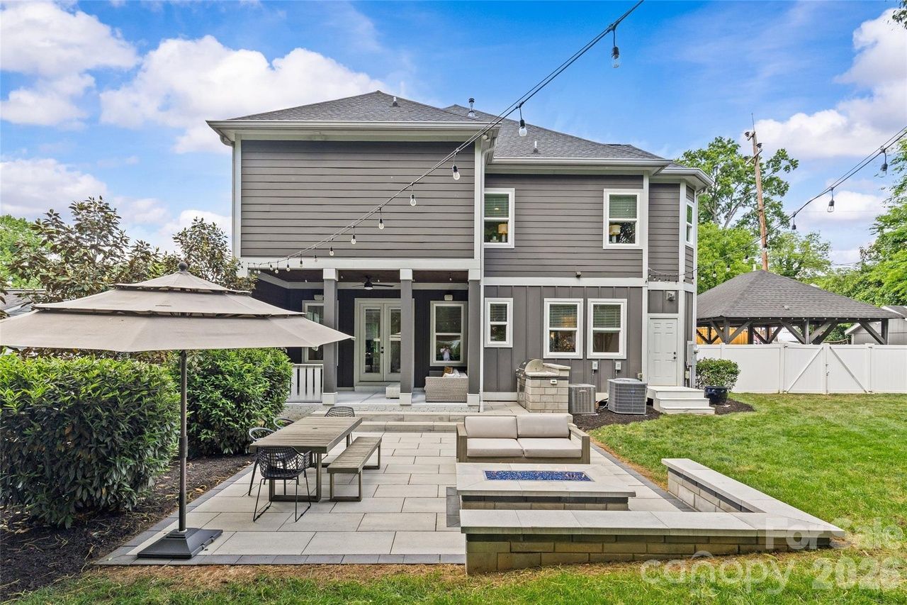 Backyard of a two-story gray house with white trim. A paved patio features a built-in fire pit, beige outdoor sofa, dining table, string lights, and a large umbrella; green lawn, hedges, and a distant gazebo.