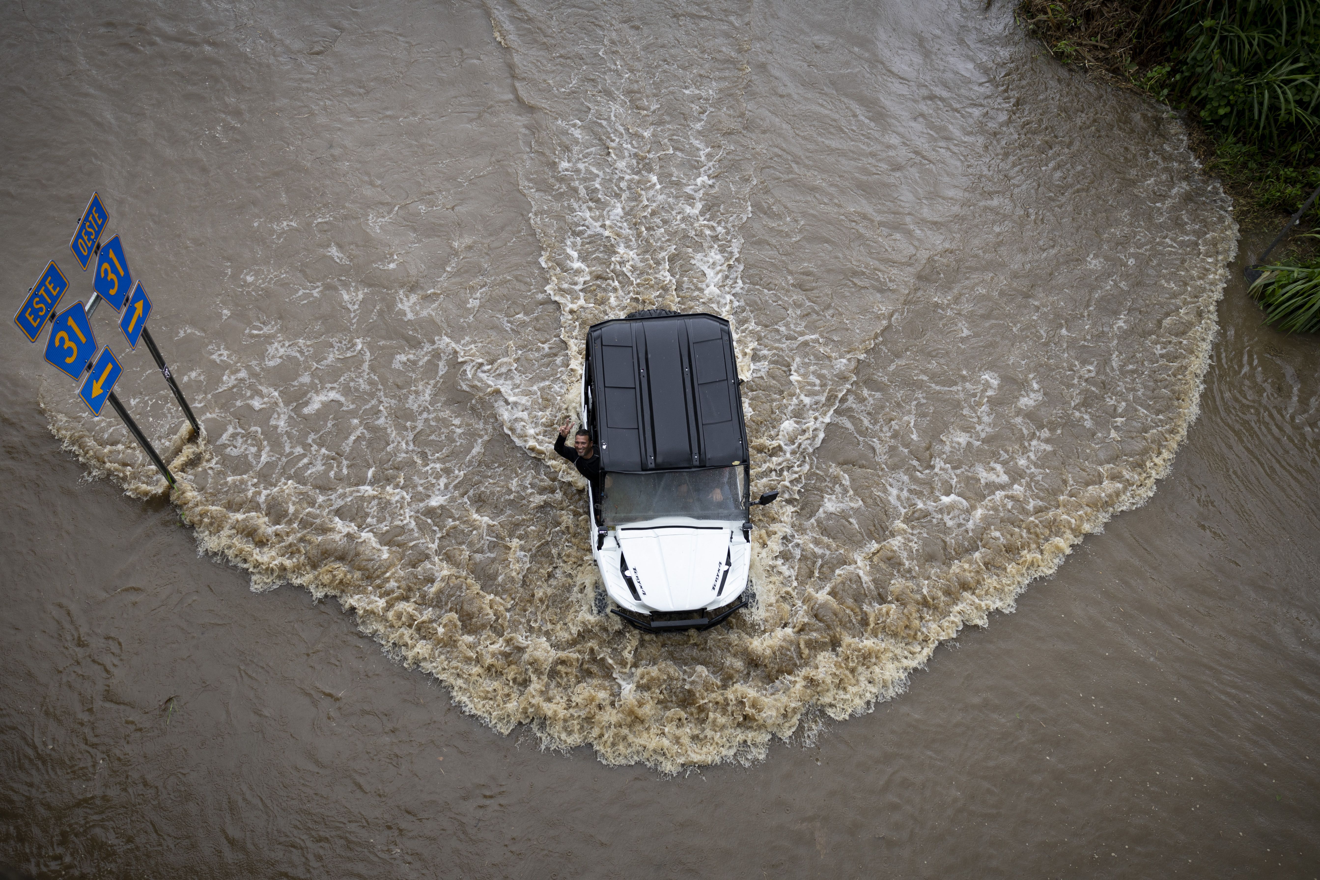 White off-road vehicle with black roof driving through deep floodwaters, creating waves; driver waving out window near street signs indicating routes 31 East and West.