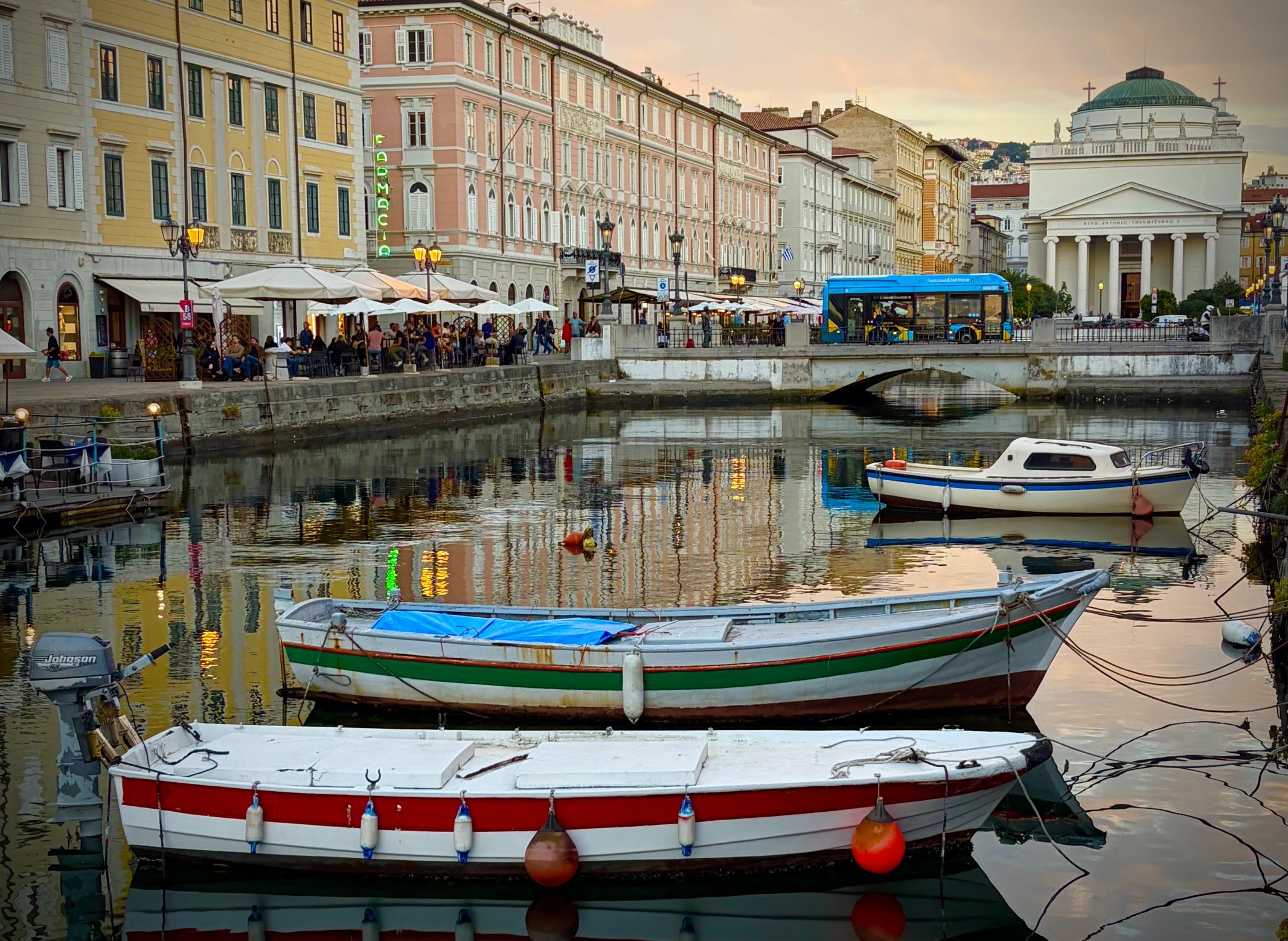 Pastel buildings line a canal with outdoor cafes under umbrellas; colorful boats docked along the water, a blue bus on a bridge, and a white domed building in the background.