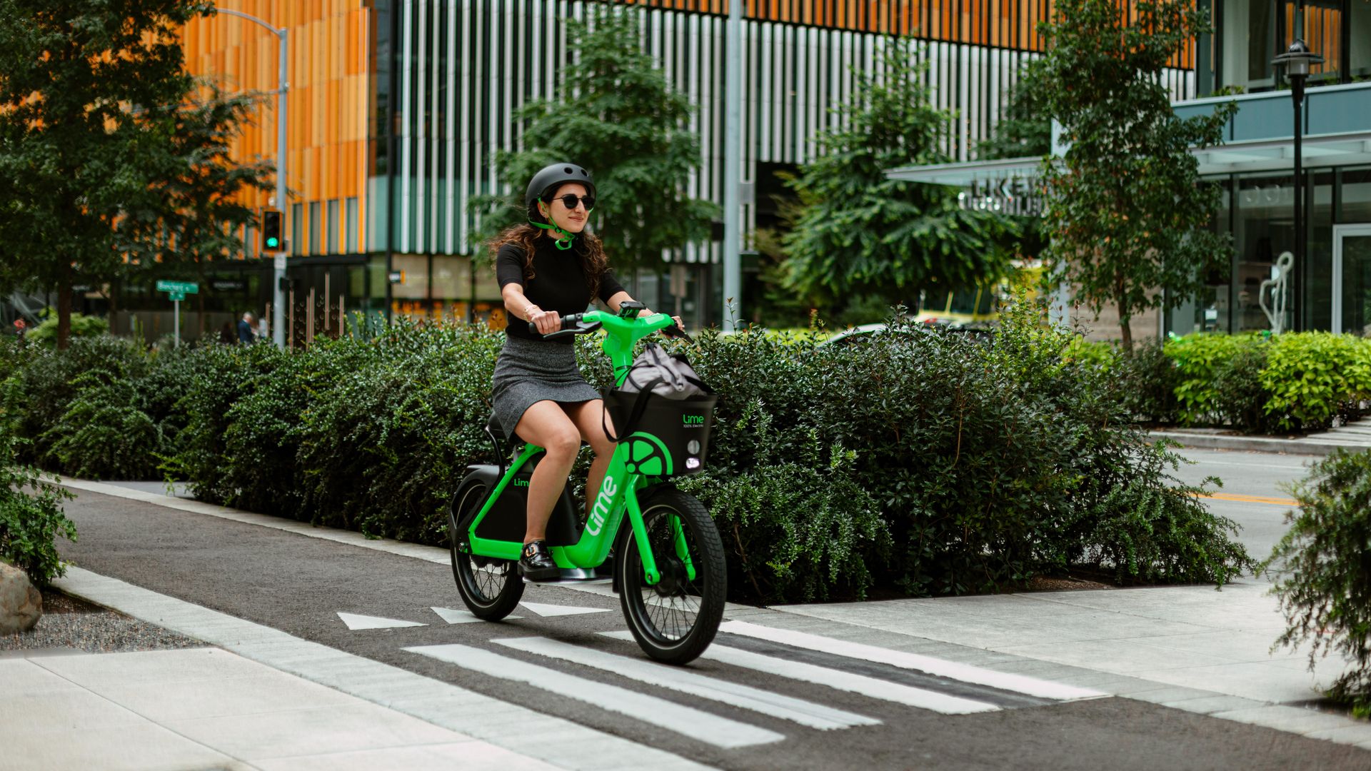 A woman wearing a black helmet and sunglasses rides a bright green Lime electric bike on a city sidewalk with green bushes and modern buildings with orange accents in the background.