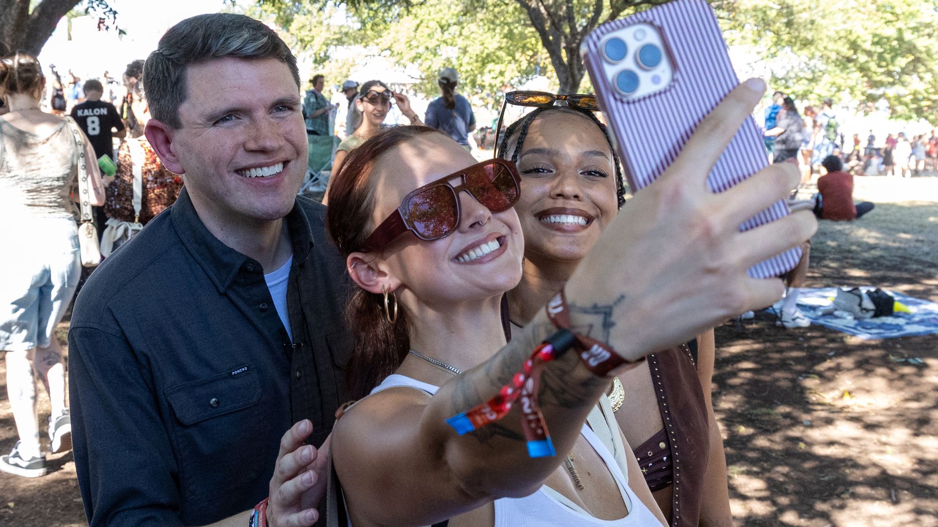 Three friends take a sunny park selfie: a man in a dark shirt on the left, two smiling women in front, one holding a purple striped phone with blue camera lenses, trees and people in the background.