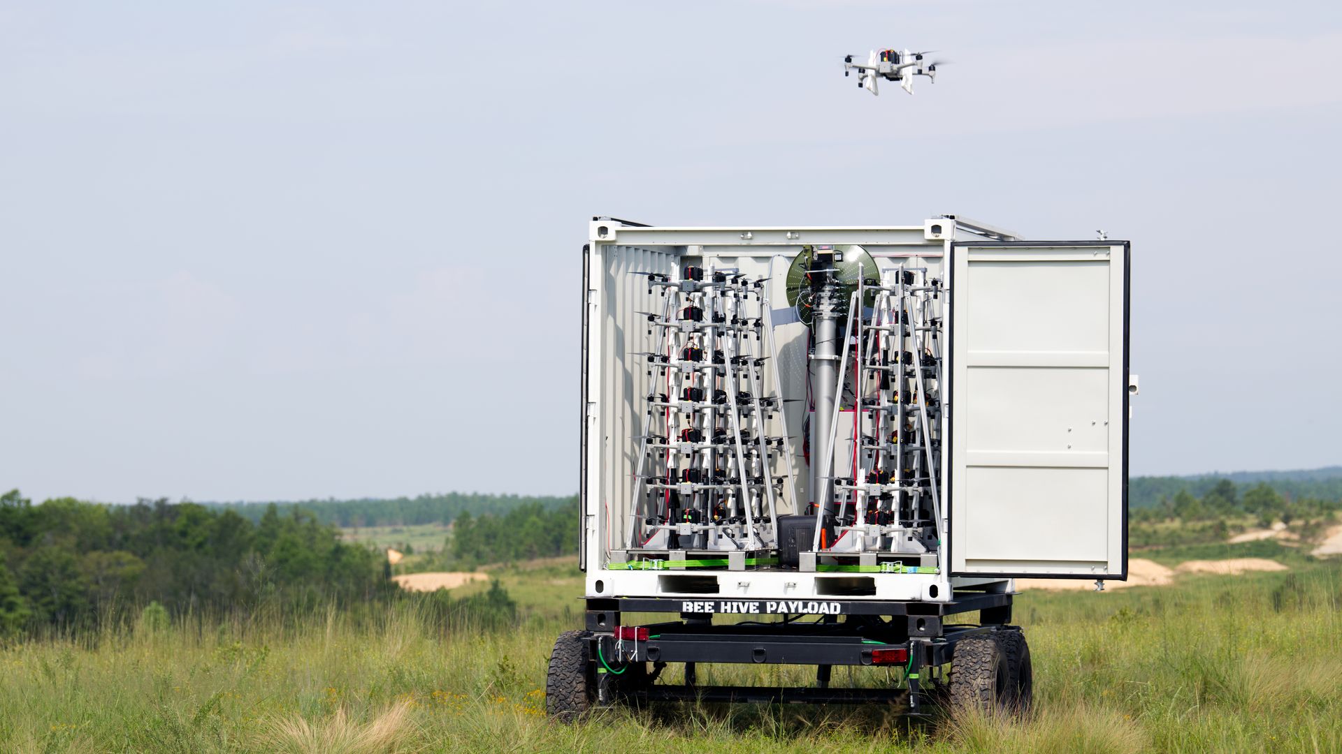 Open container on a trailer filled with drones. A white quadcopter hovers in a clear blue sky.