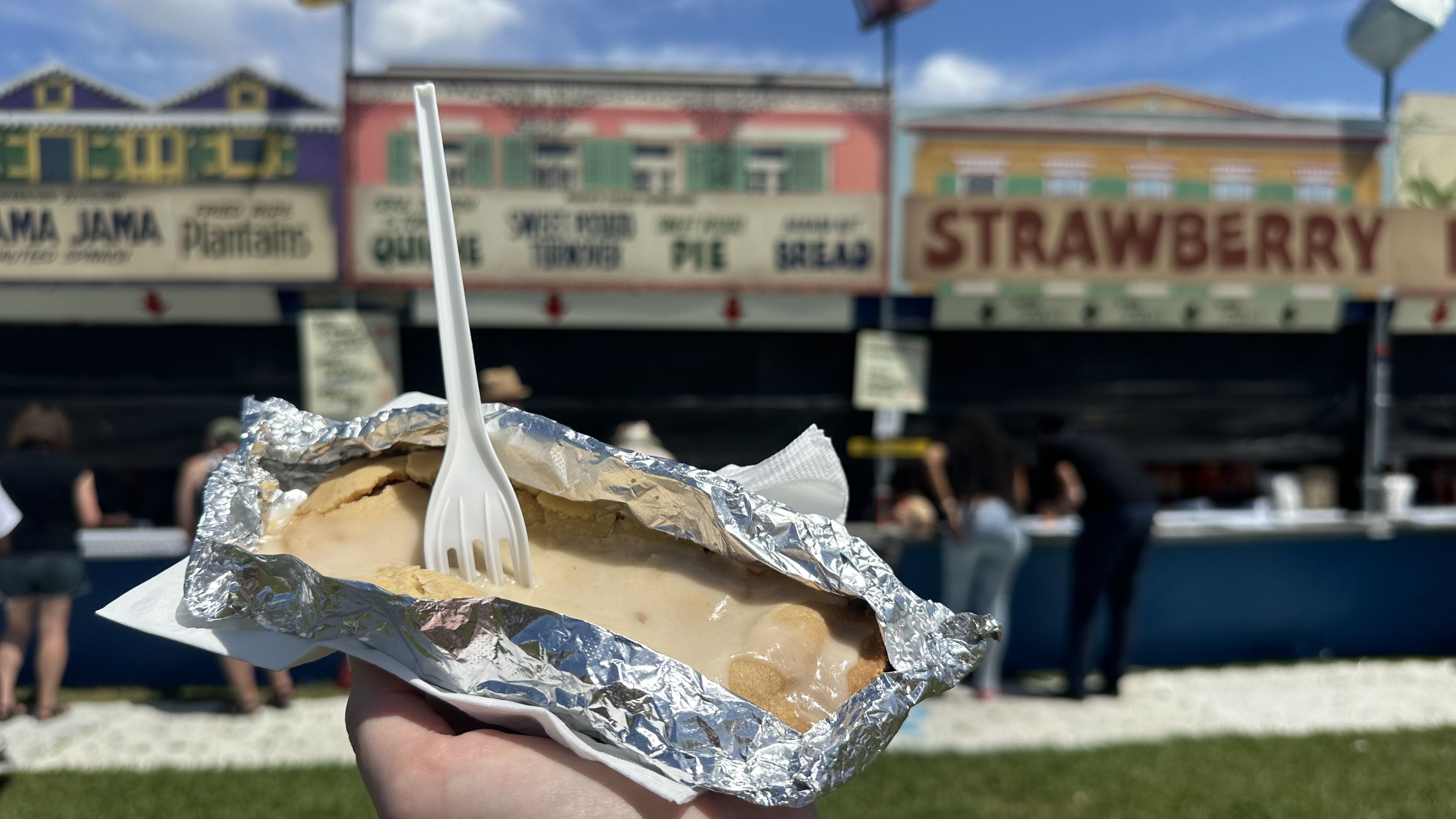 A close-up image of a sweet potato turnover, covered in icing with a plastic fork sticking out of it. Jazz Fest vendors' booths are seen in the background.