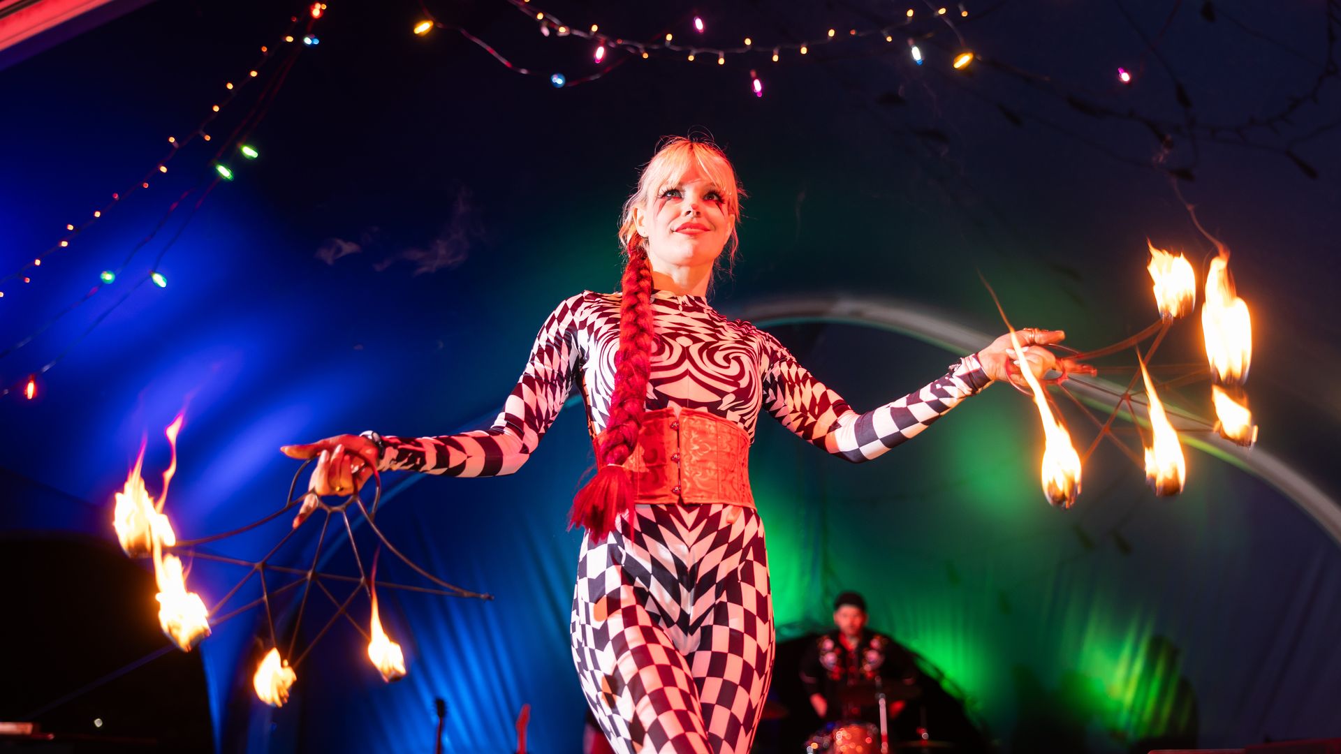 Female fire performer with a long red braid, wearing a black-and-white geometric outfit and red corset, spins fire torches on stage beneath colorful string lights.