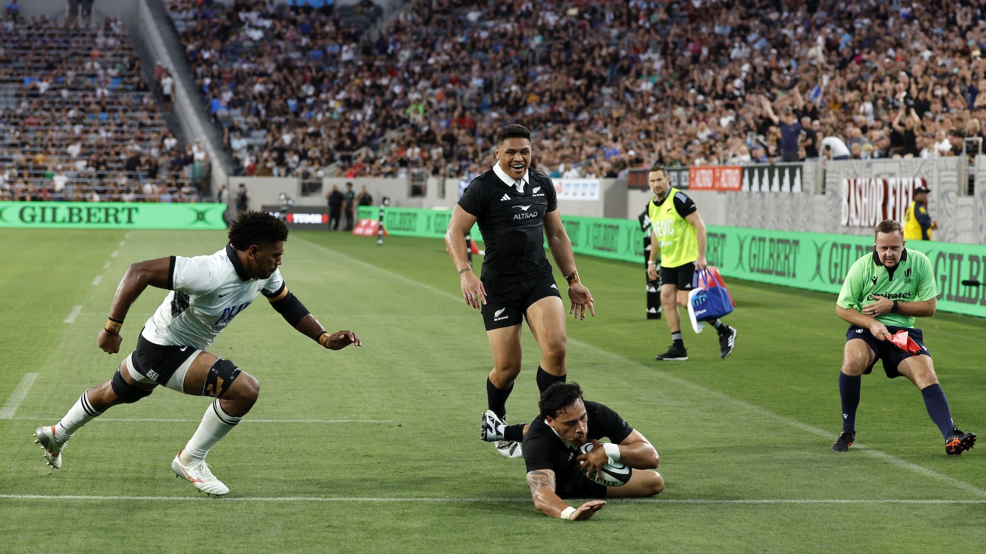 A rugby player dives on the field to score as a defender runs toward him and a teammate smiles behind him during a match at Snapdragon Stadium.