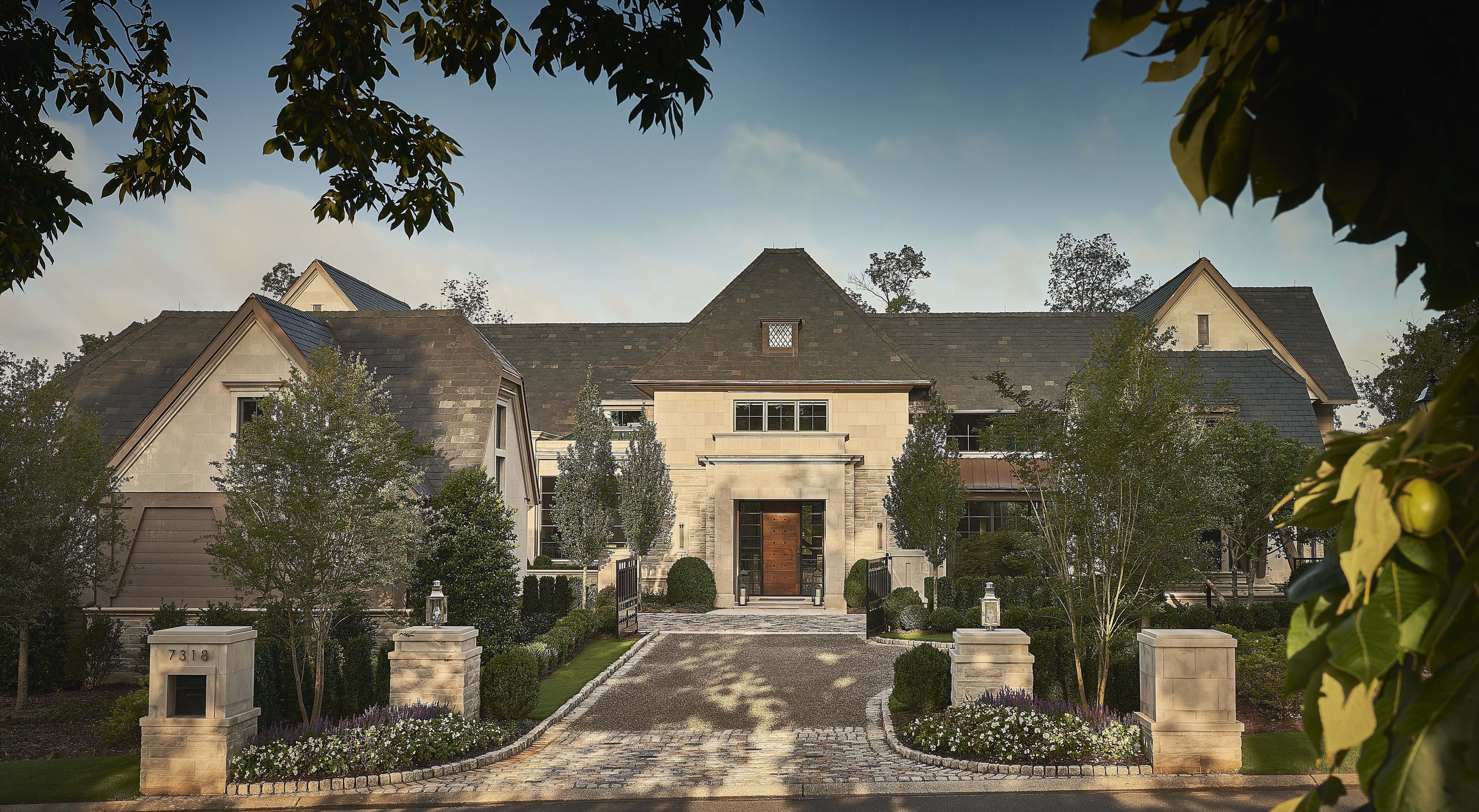 Frontal view of a large stone mansion with a slate roof, manicured shrubs, and a cobblestone driveway leading to a wooden front door; trees frame the blue sky.