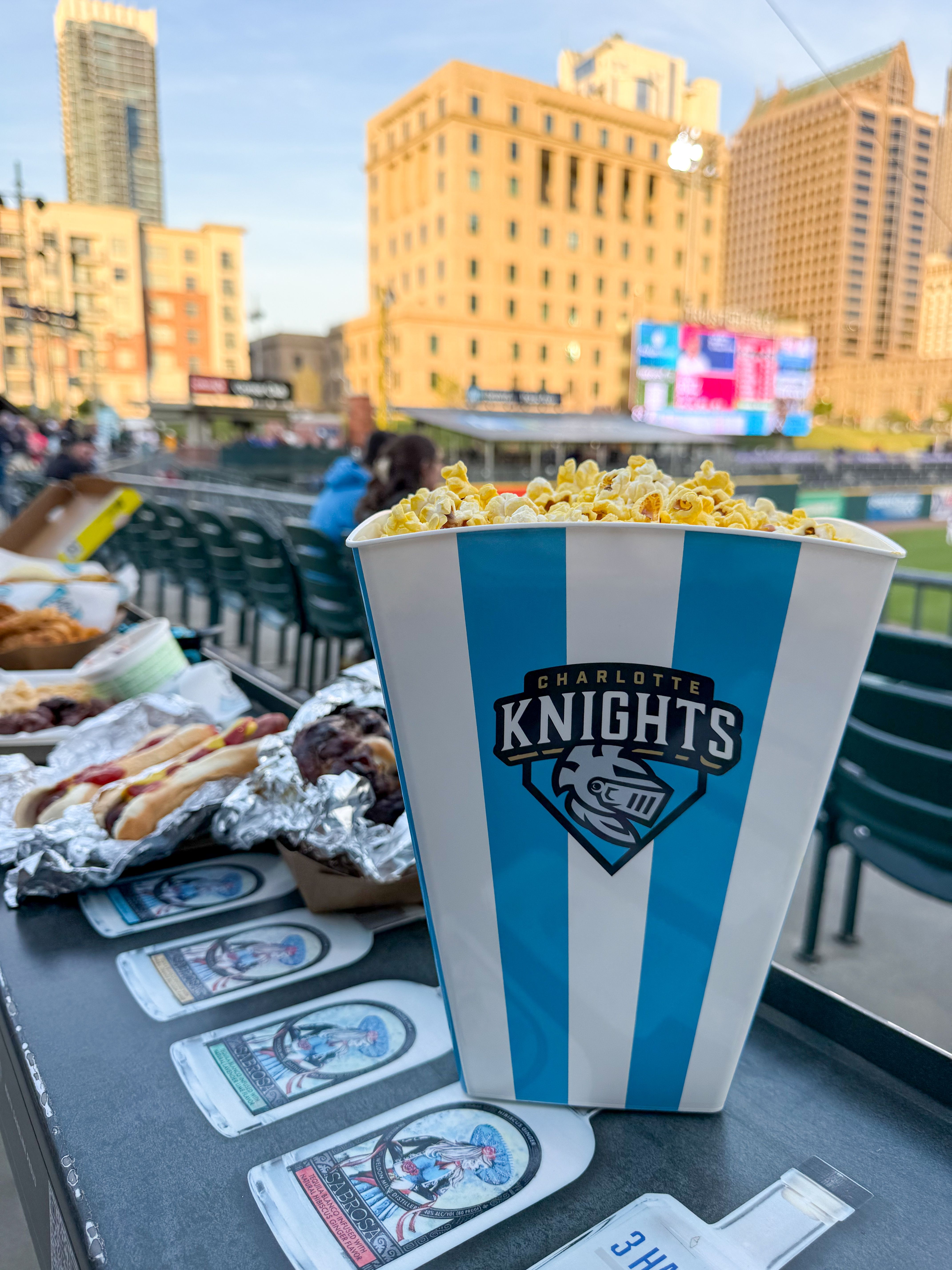 Blue and white striped popcorn tub with the Charlotte Knights logo brims with popcorn on a table at a baseball stadium, near foil-wrapped hot dogs, with city buildings in the background.