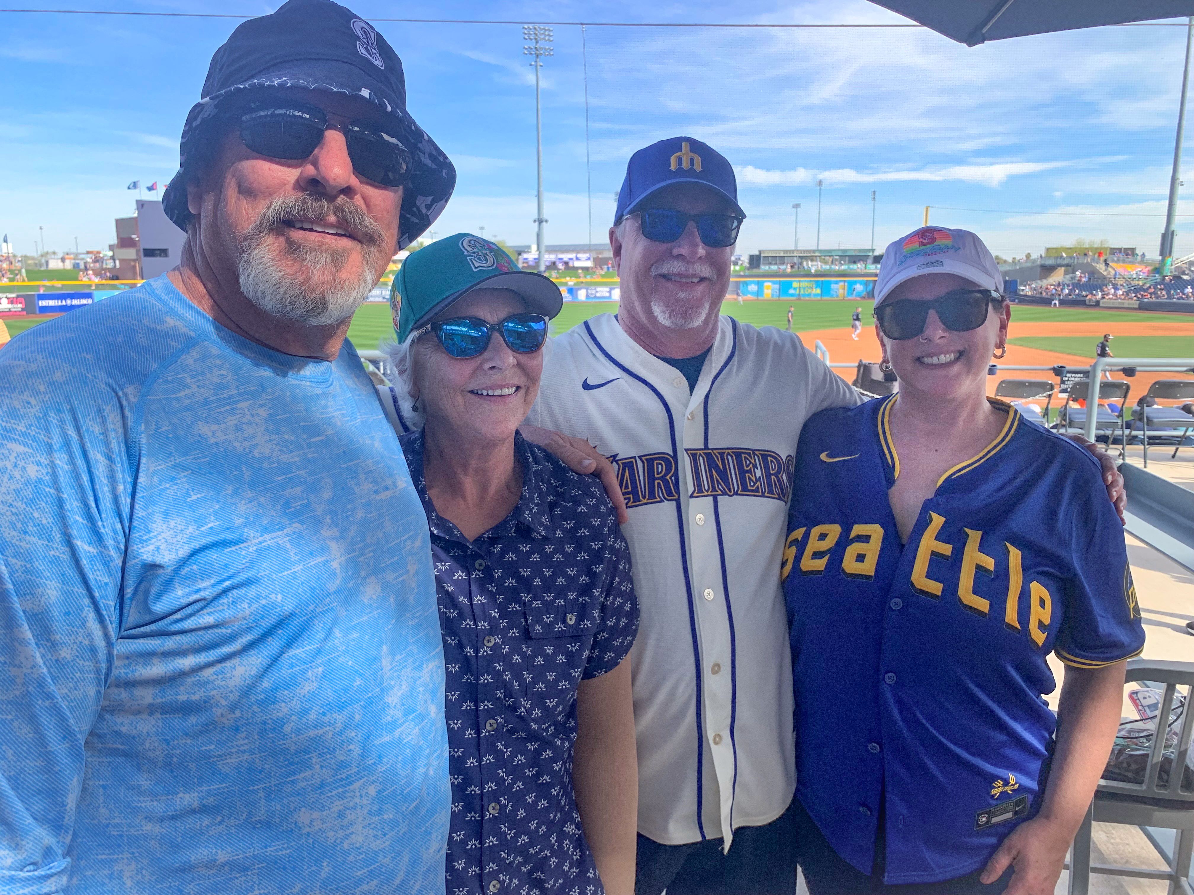 Four people stand in front of a ball park wearing hats and sunglasses, two of them wear Mariners or Seattle jerseys.