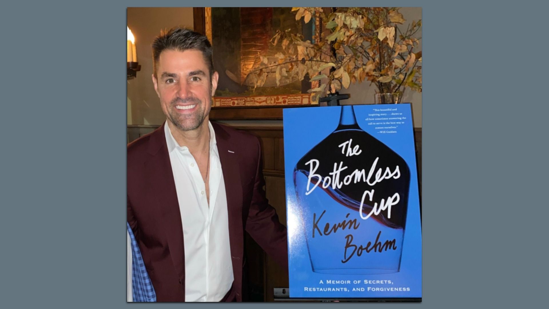 A smiling man in a white shirt and maroon jacket stands beside a blue poster for the book "The Bottomless Cup" by Kevin Boehm, subtitled "A Memoir of Secrets, Restaurants, and Forgiveness."