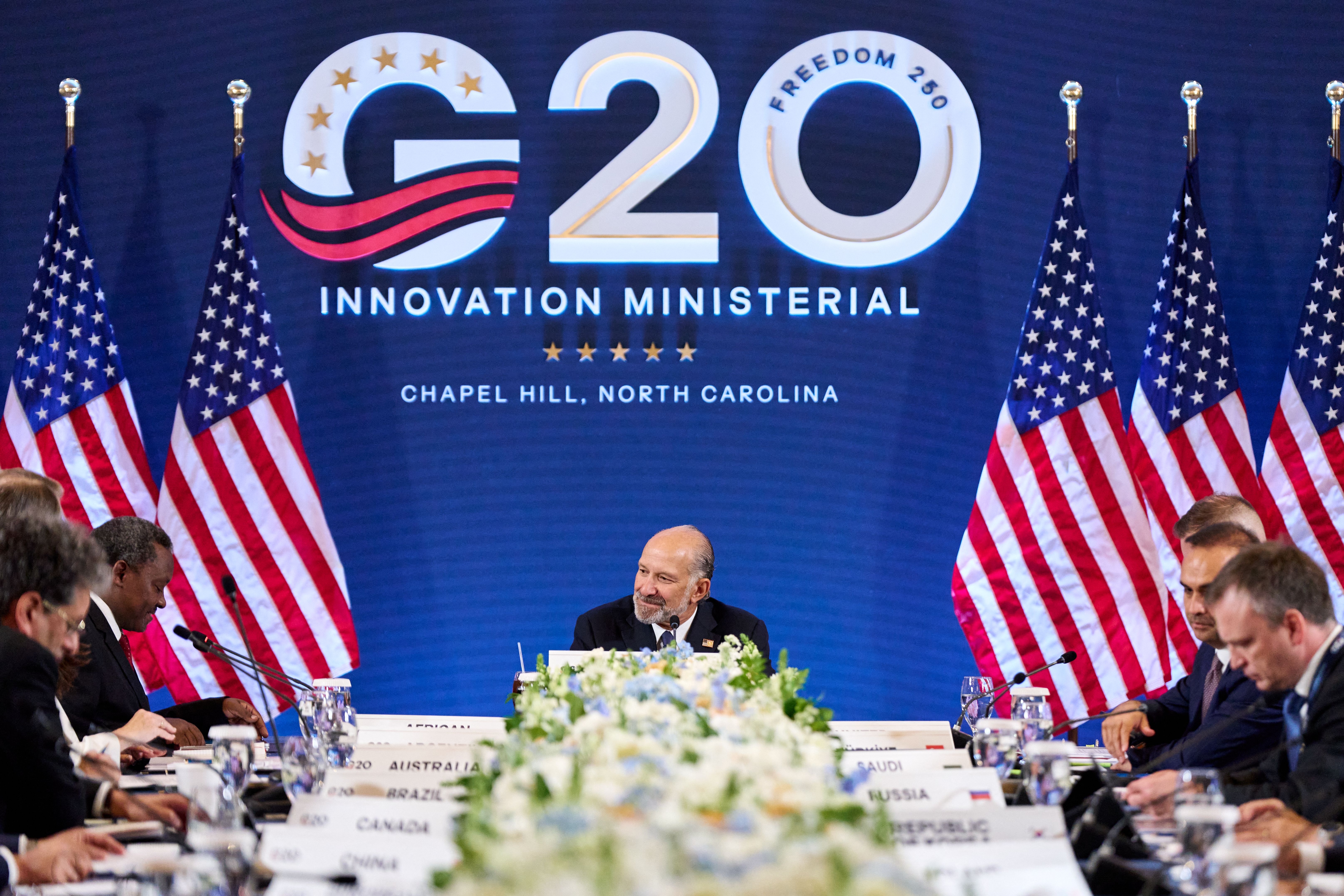Commerce Secretary Howard Lutnick, a bald man with a white beard, sits at a table with multiple men in suits. The blue wall behind him is lined with American flags and reads "G20 INNOVATION MINISTERIAL."