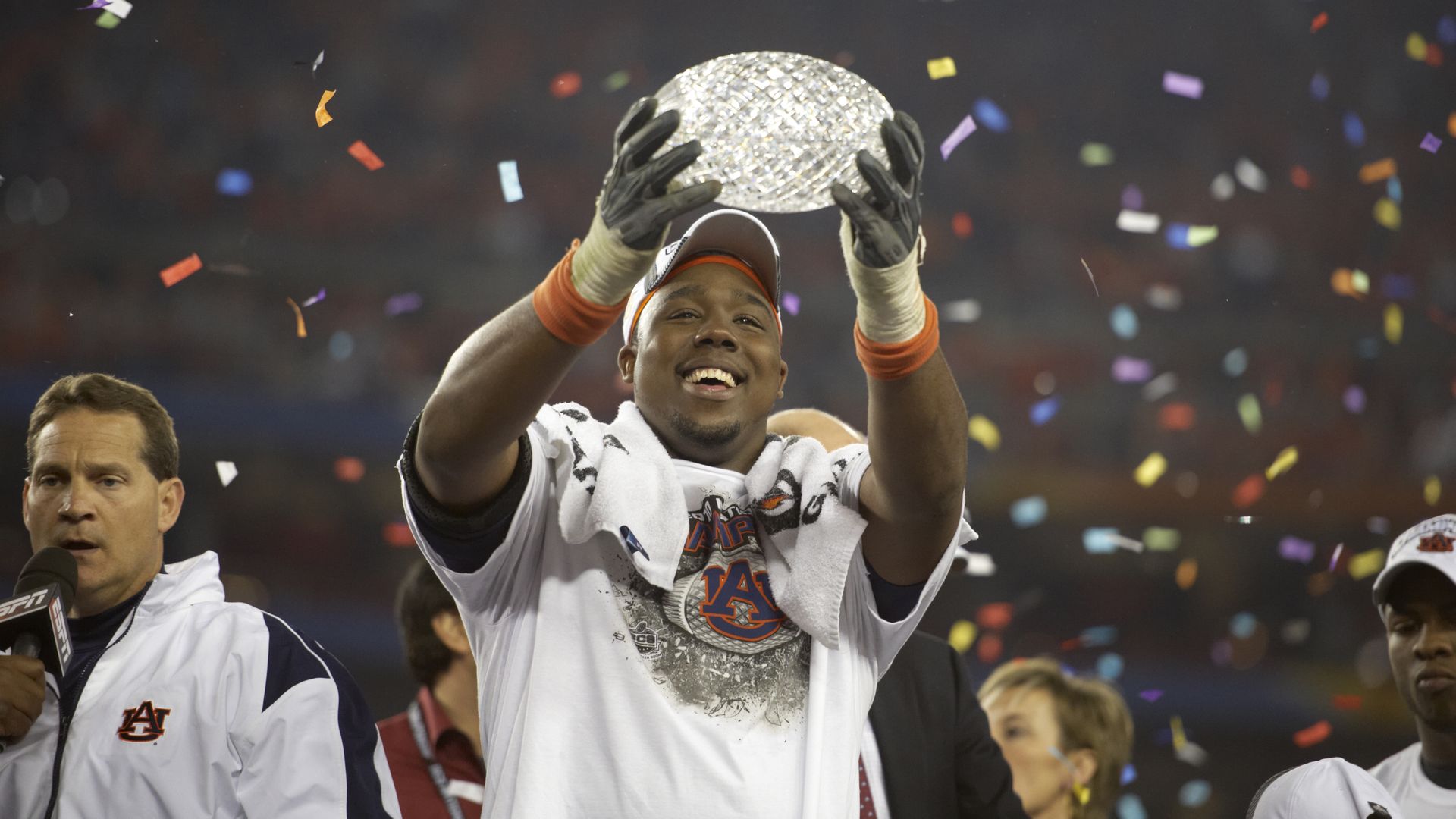 Smiling Auburn football player holding a crystal trophy aloft amid falling colorful confetti, celebrating a championship victory with teammates and coaches nearby.