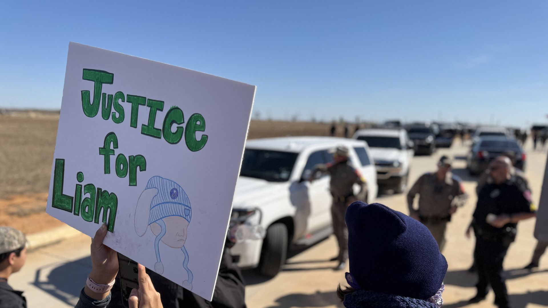A person holds a sign that says "Justice for Liam" in green letters with a drawing of a child wearing a blue bunny hat.