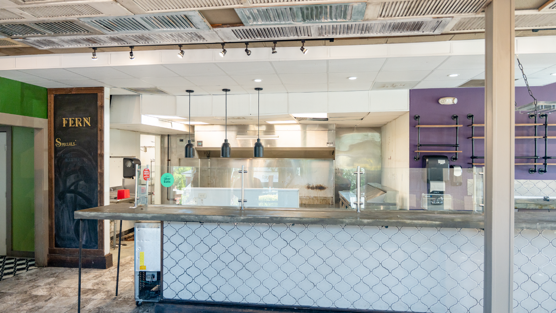 Empty restaurant kitchen with white tiled counter, black hanging lights, purple and green walls, a chalkboard with "FERN Specials," and an Uber Eats sticker on the glass partition.