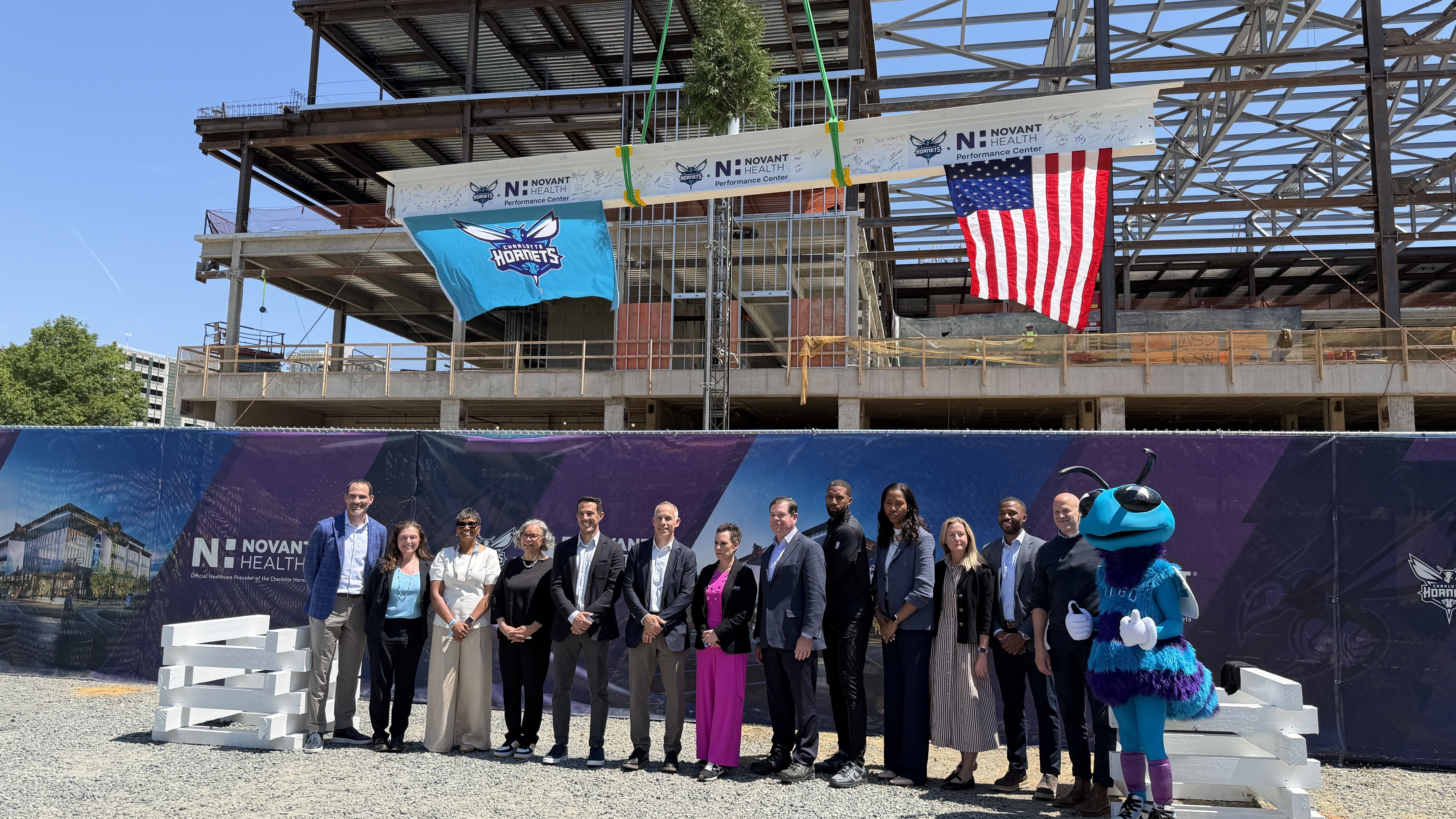 Group of professionally dressed people posing in front of a built steel-frame construction site. An American flag and a Charlotte Hornets banner hang overhead, with a blue mascot on the right.