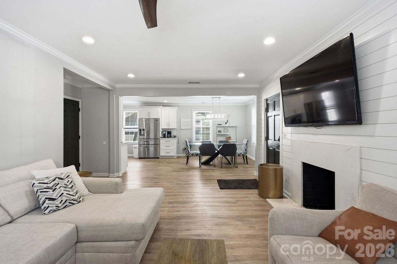 Open-concept living room with light gray walls and wood floors. A gray sectional faces a wall-mounted TV above a white fireplace; kitchen and dining area visible in the background.