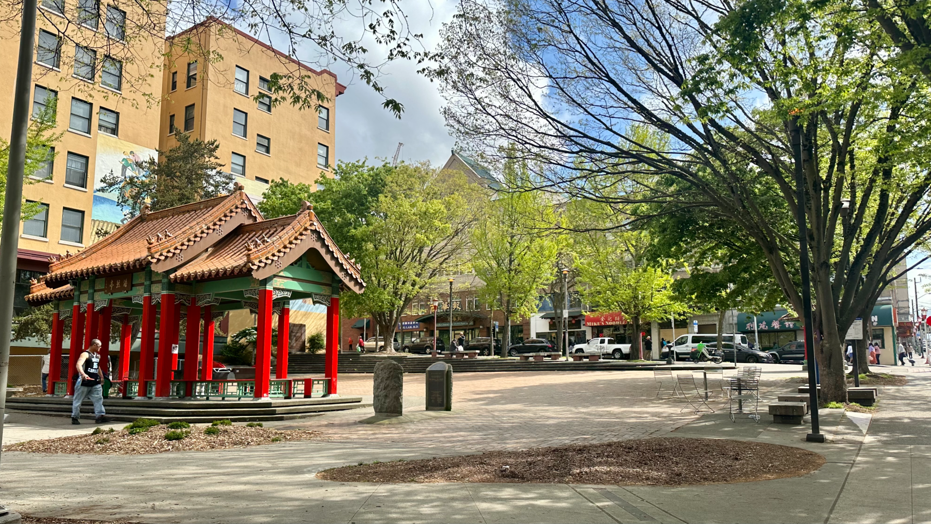 The Grand Pavilion with its red columns and pointed roofs is shown in Hing Hay Park, along with trees and an open plaza.