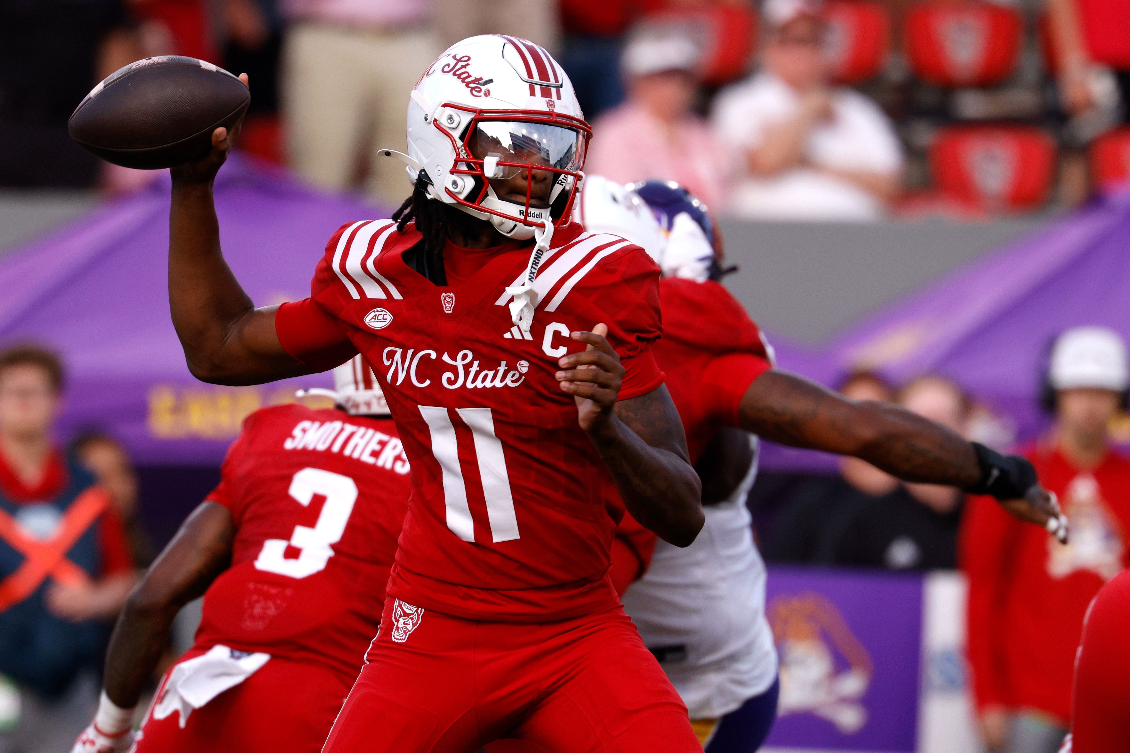 NC State football quarterback in red uniform #11 about to throw a football during a game, with a teammate and opponents in background.