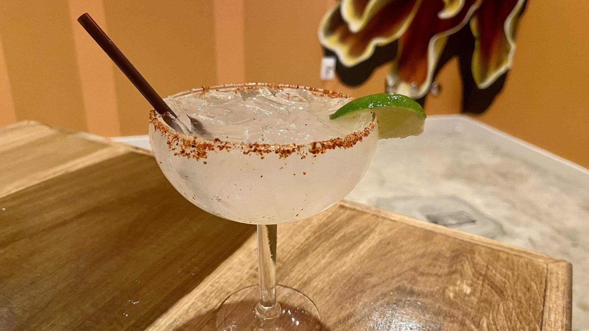 A close-up of a margarita, rimmed with tajin and a lime wedge, in a stemmed glass with a brown straw, sitting on a wooden table. An orange wall and tropical plant decor are blurred in the background at Tres Arcos at Mexico Ceaty.