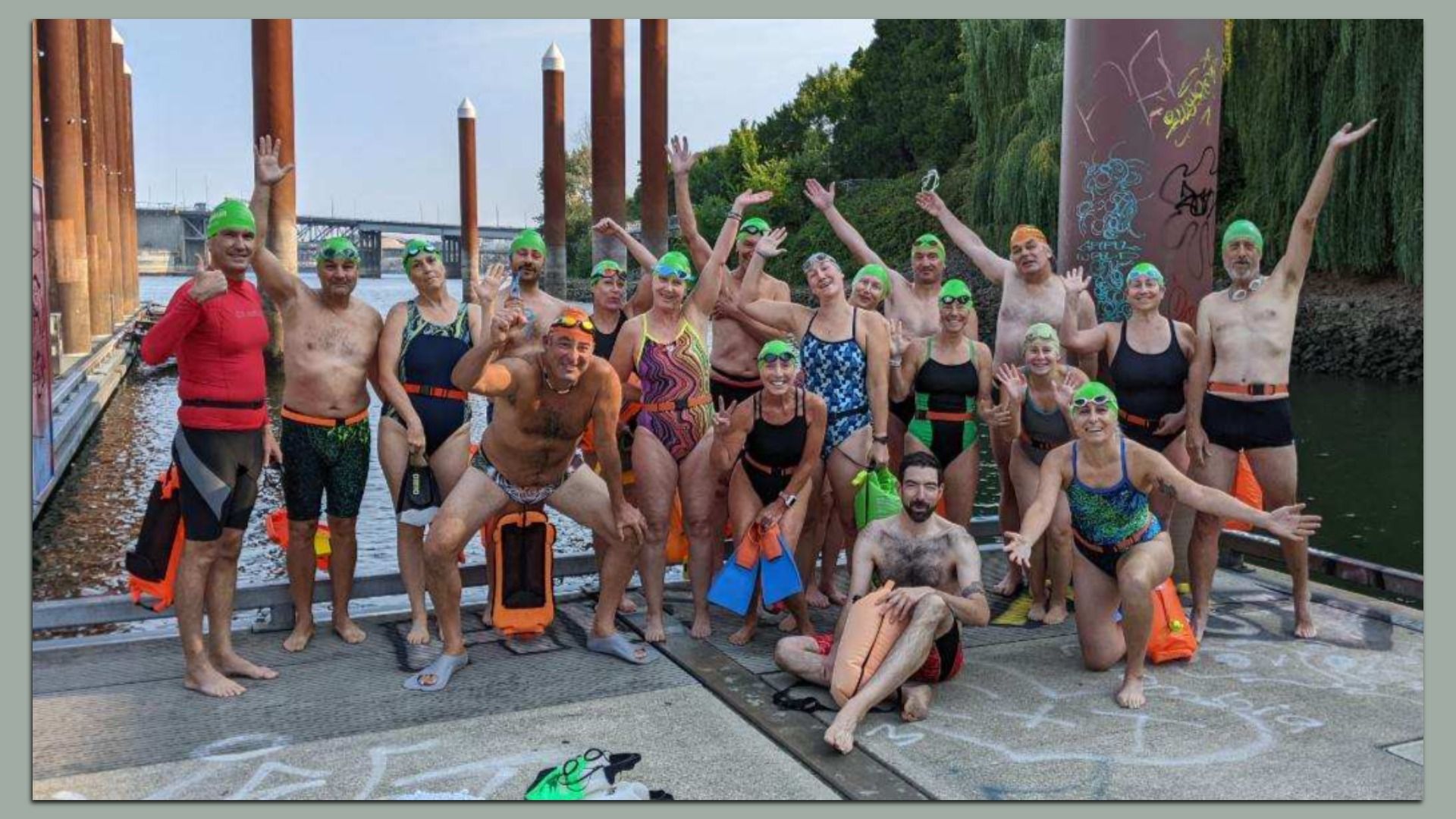 A group of open water swimmers, wearing colorful swimsuits and bright green swim caps, pose and smile enthusiastically on a dock by the river. 