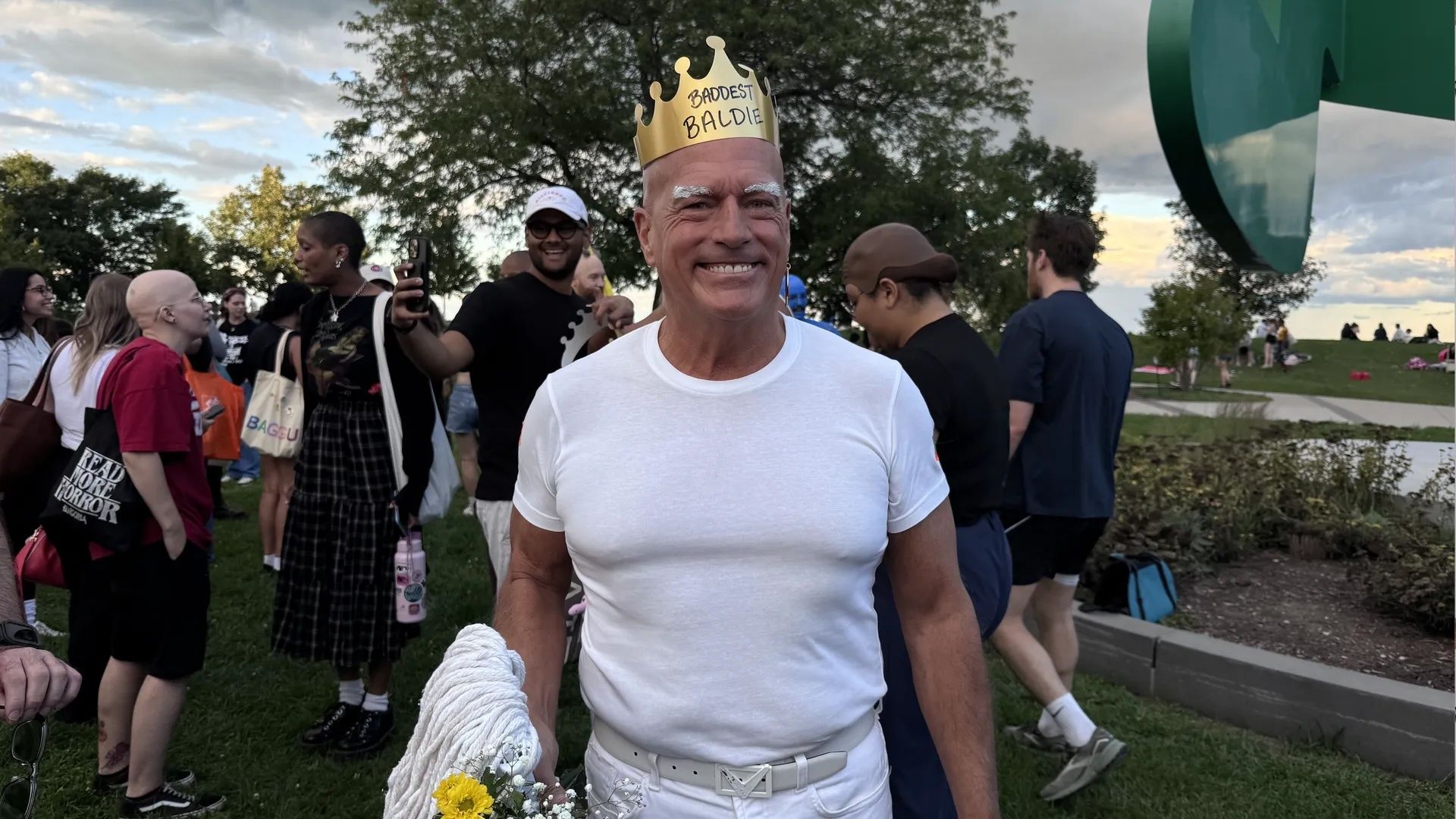 Smiling bald man in white t-shirt and pants wears gold crown saying "Baddest Baldie" at outdoor gathering with people and trees in background.