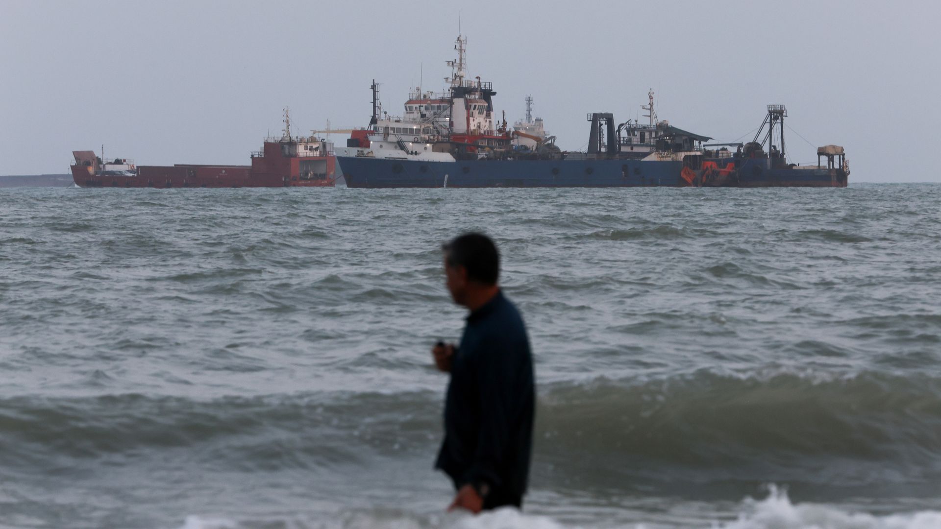 A blue and white cargo ship sits among several smaller red and gray vessels on a choppy sea, with a blurred person in the foreground and a hazy gray sky.