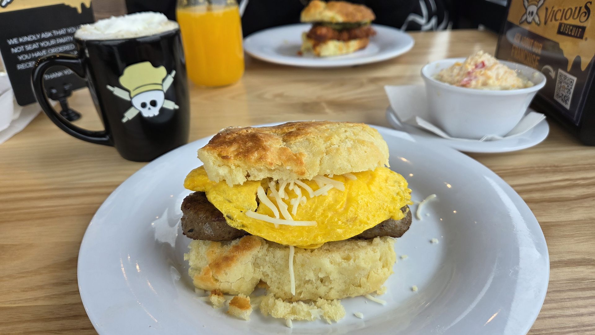 Breakfast biscuit sandwich with scrambled egg, sausage patty, and shredded cheese on a white plate. Black mug with skull logo, orange juice, and side of creamy salad on wooden table.