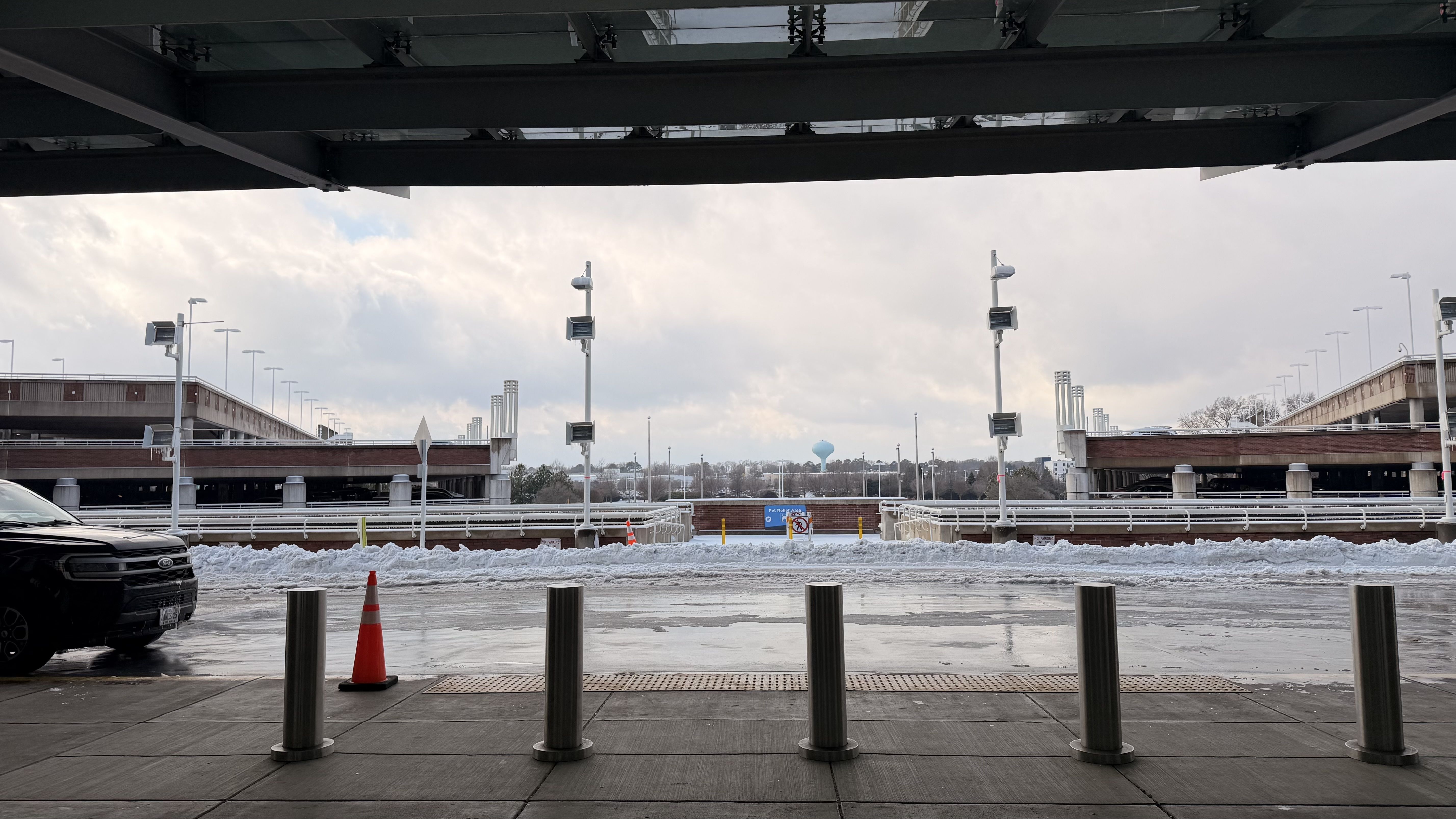 View from under a covered area showing a snowy parking lot and two multi-level parking structures with metal poles and a traffic cone in the foreground under a cloudy sky.