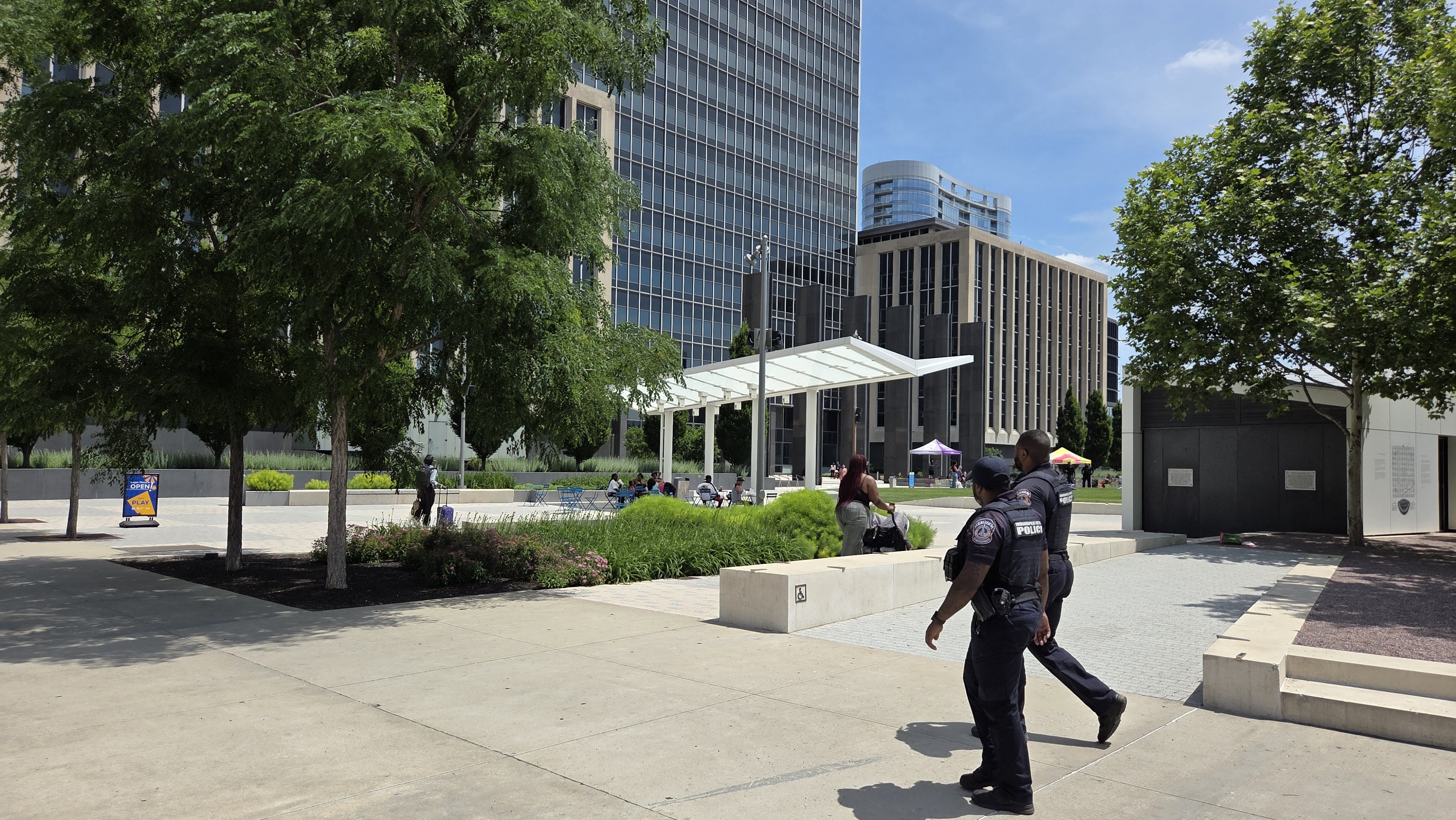 Sunny urban plaza with tall glass towers, green trees, and a white canopy. People sit at tables while police officers walk in the foreground.