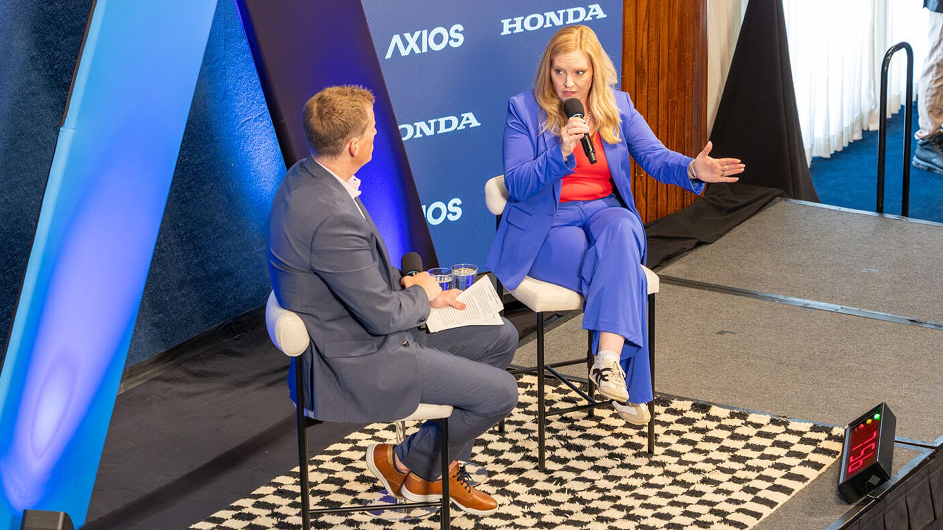 Two people in a staged interview: a man in a gray suit and a woman in a blue suit with a red top, speaking into microphones. A blue AXIOS/HONDA backdrop and a blue decorative panel on the left.