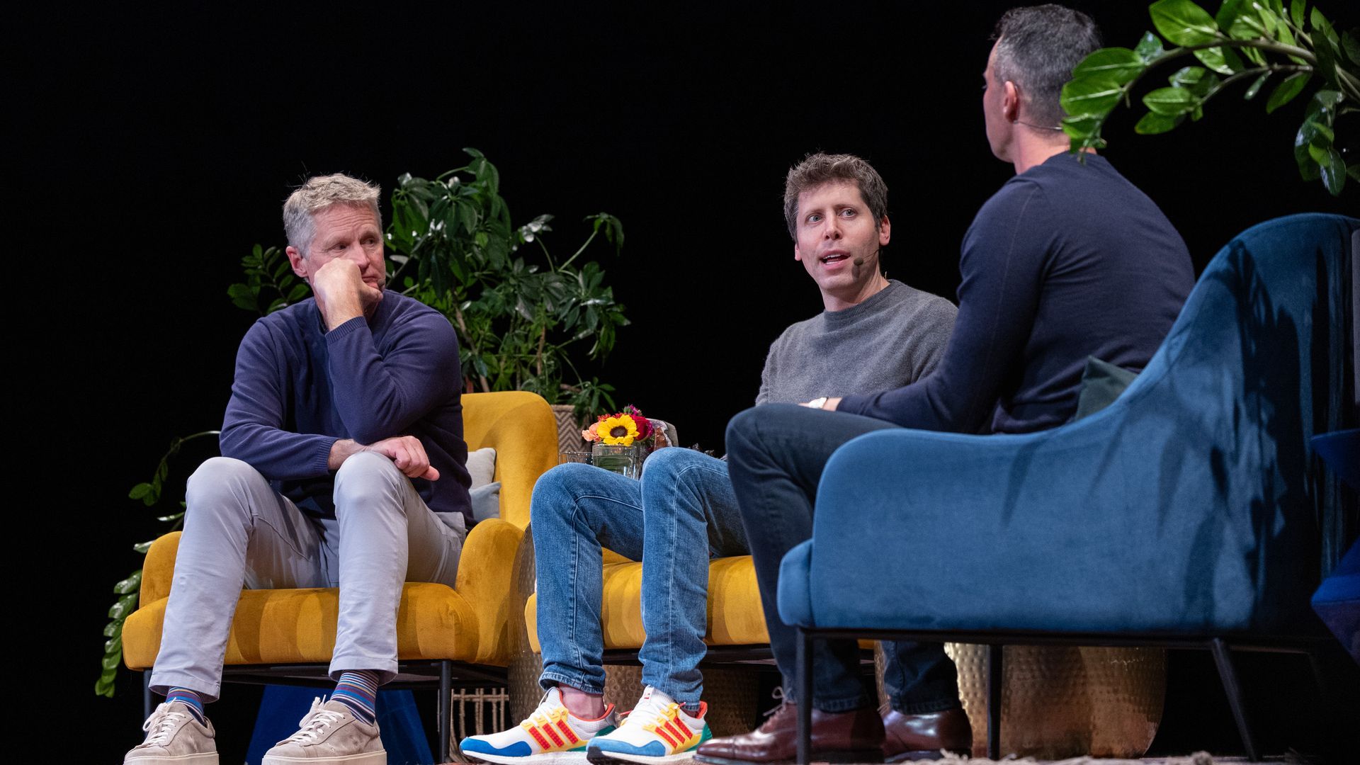 Three men seated on yellow and blue armchairs on a stage with plants behind them, two wear microphones, one man gestures while speaking, casual clothing and sneakers visible.