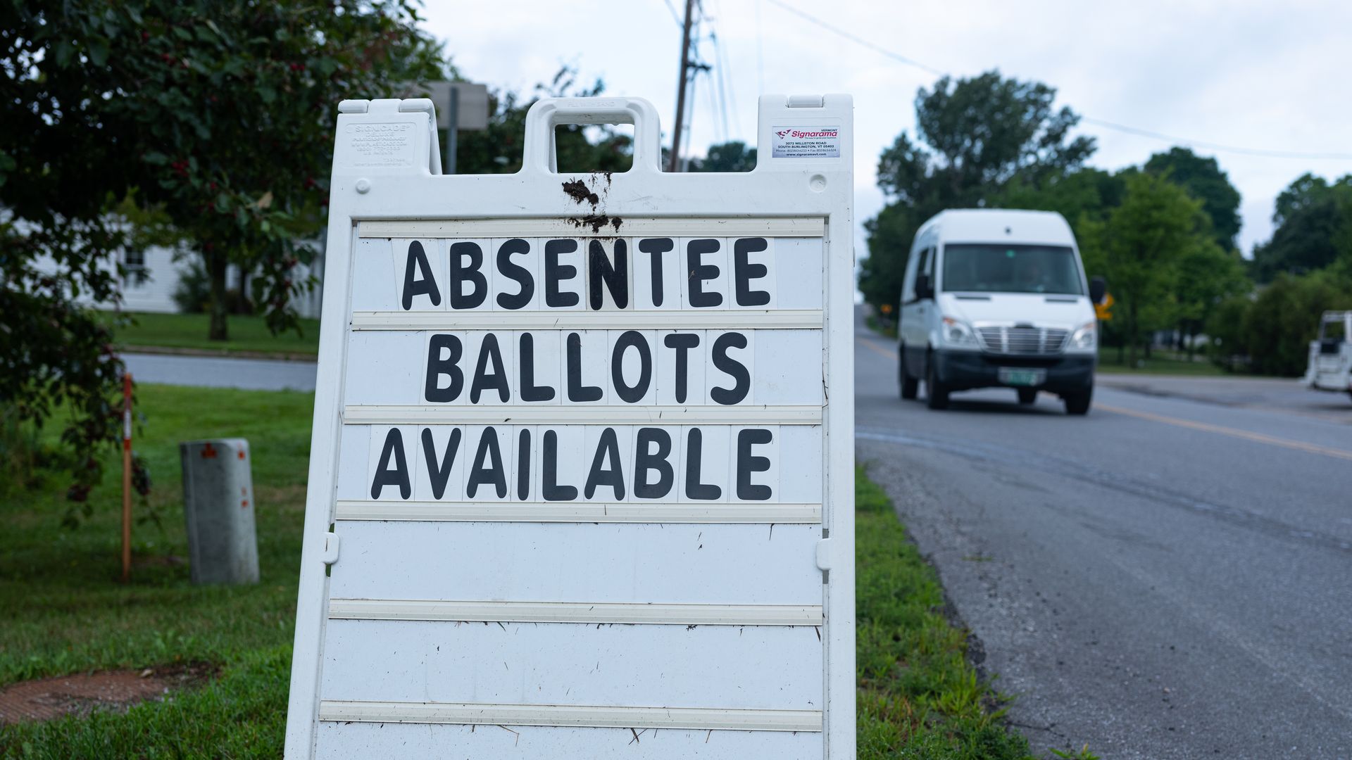 Photo of a sign that says "absentee ballots available" and sits on a bed of grass next to a road