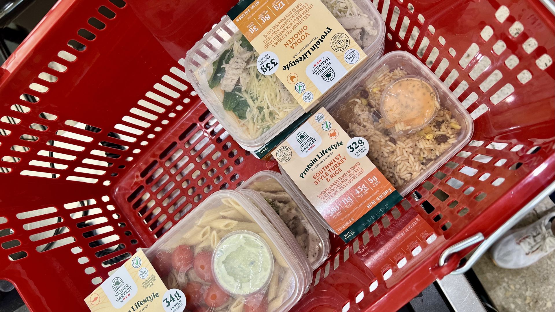 Red shopping basket with three Protein Lifestyle packaged meals: Green Goddess Salmon, Southwest Style Turkey & Rice, and Vodka Chicken, each showing nutrition and protein info.