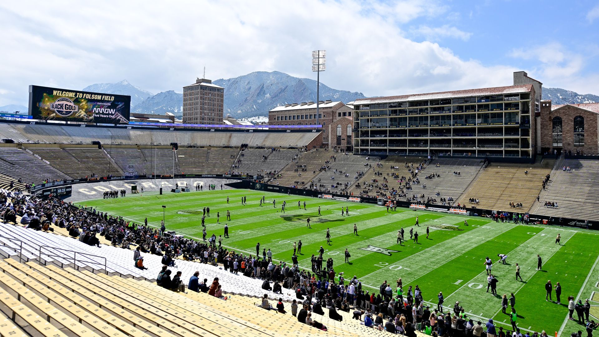Bright sunny day at Folsom Field with green field and scattered spectators. Mountains and blue sky with clouds in background. Large scoreboard reads "Welcome to Folsom Field."