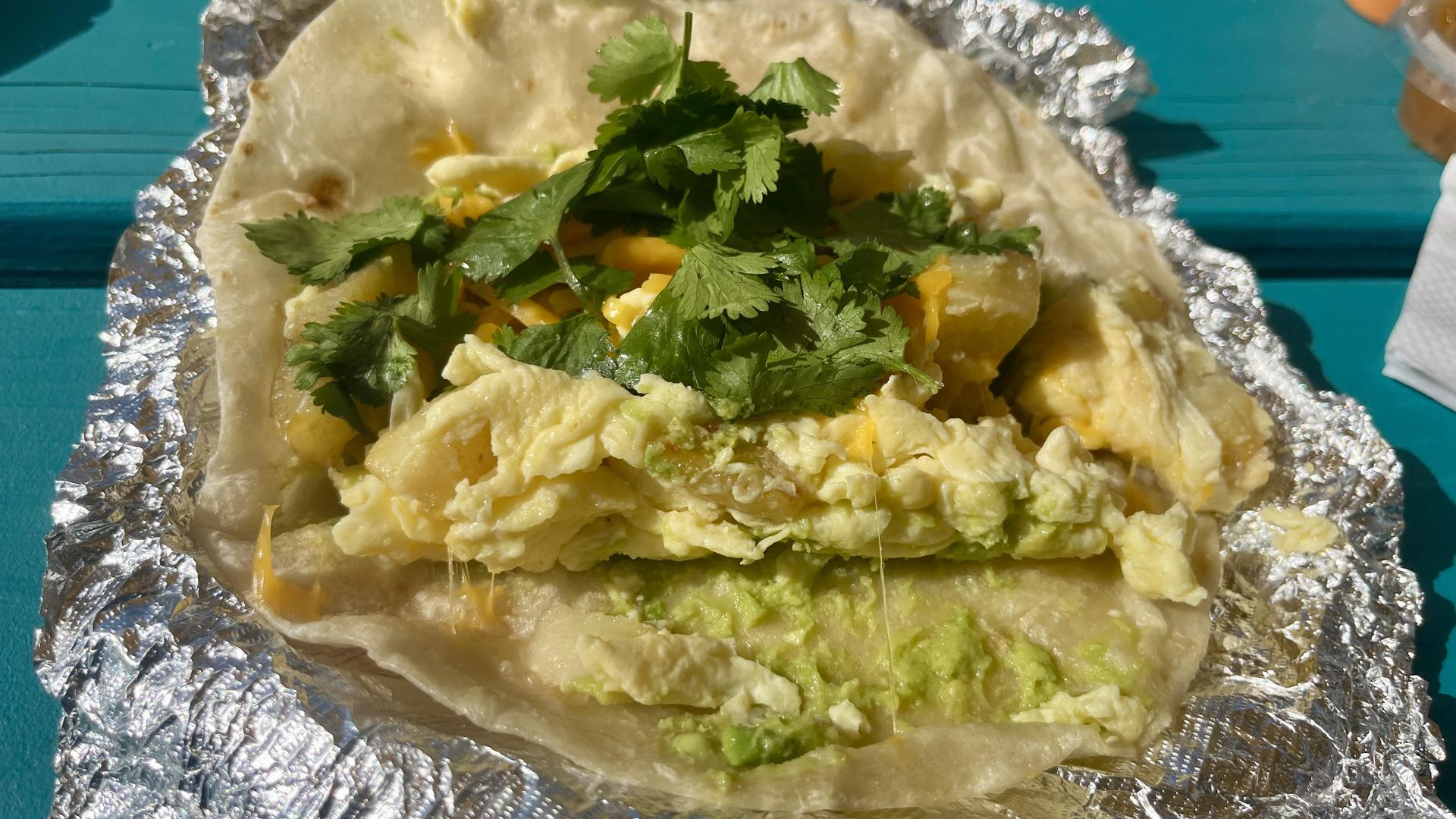 An egg and potato breakfast taco on top of foil on a blue table.