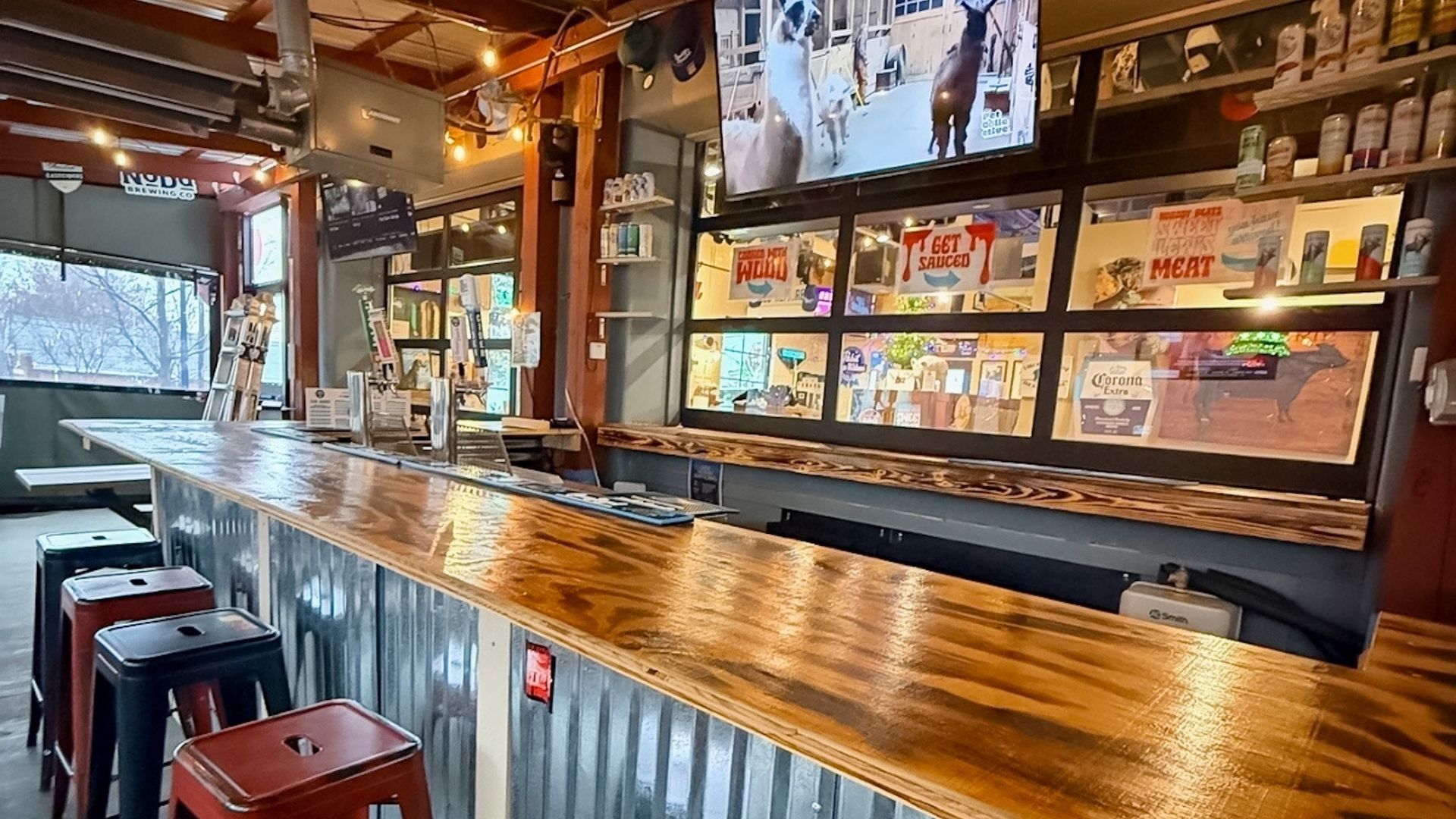 Empty bar with corrugated metal front and polished wooden countertop, black and red metal stools, TV showing llamas, various drink signs including Corona Extra and "Get Sauced."