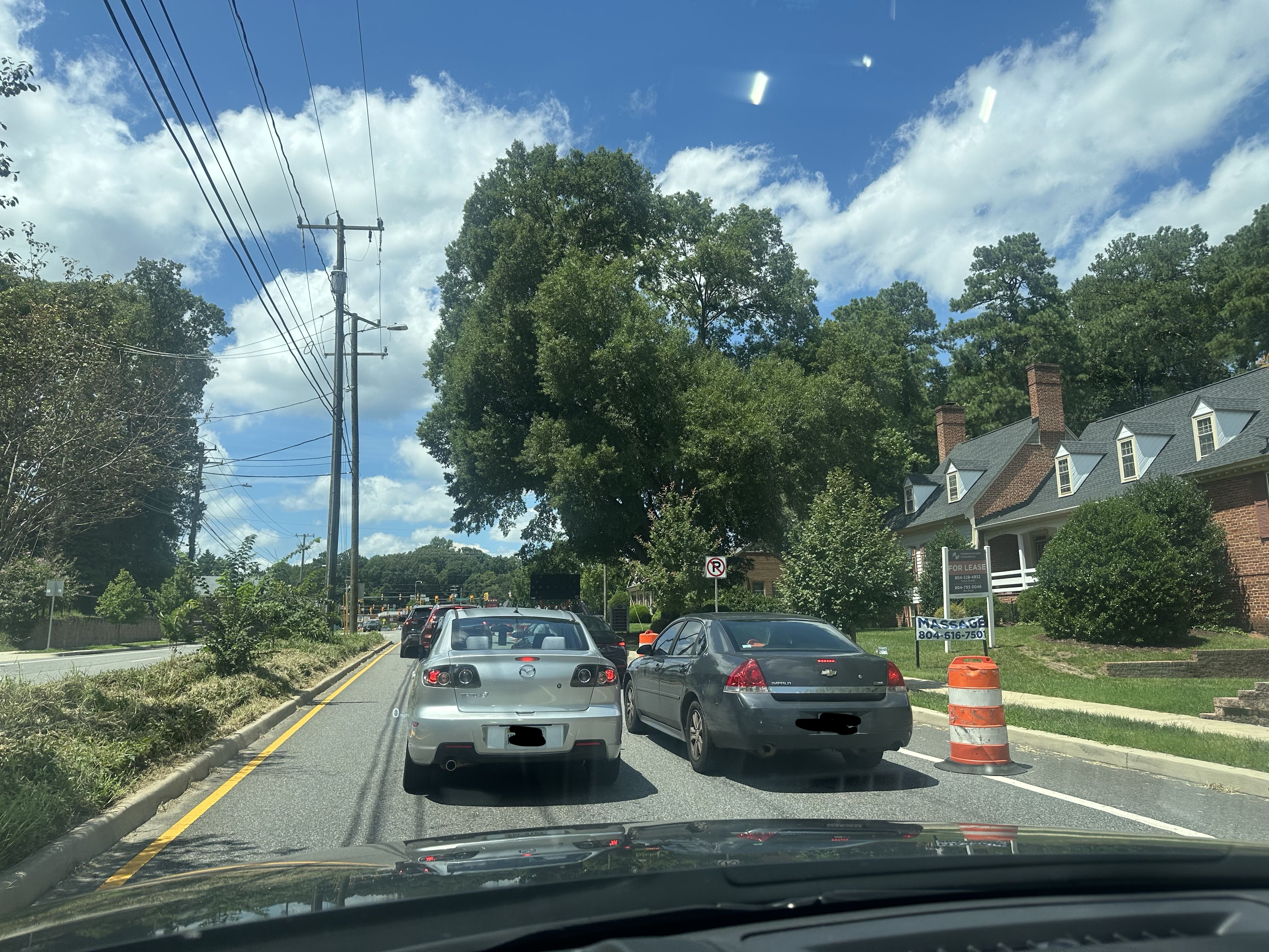 View from a car on a street: cars in traffic, tall trees along the curb, overhead power lines, brick houses with chimneys on the right, a blue sky with clouds, and an orange barrel on curb.