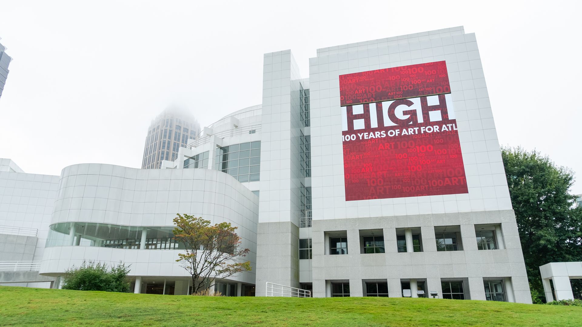 Modern white museum building with a large red panel that says HIGH and 100 YEARS OF ART, set against a cloudy sky; green lawn in the foreground and trees on the right.