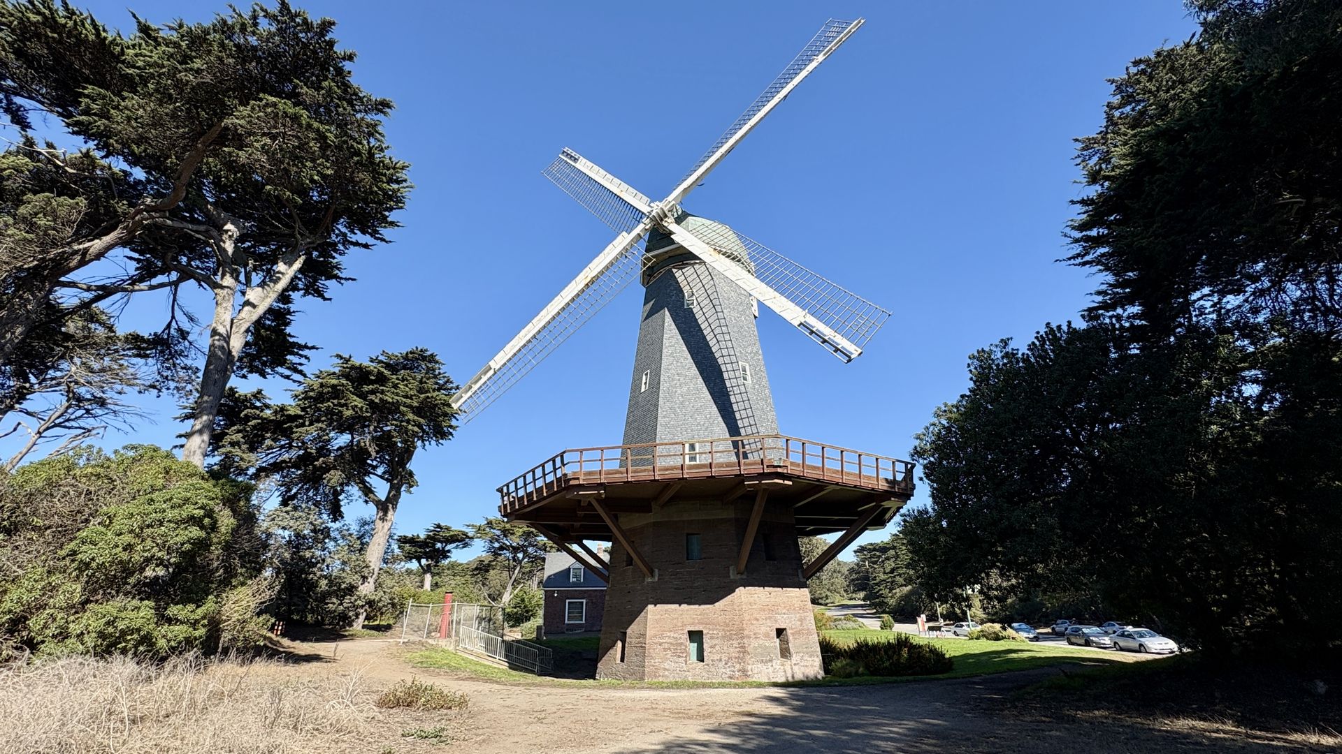 Sunlit windmill with a brick base and white lattice blades forming an X, set against a clear blue sky, surrounded by trees and a wooden observation deck around the base.