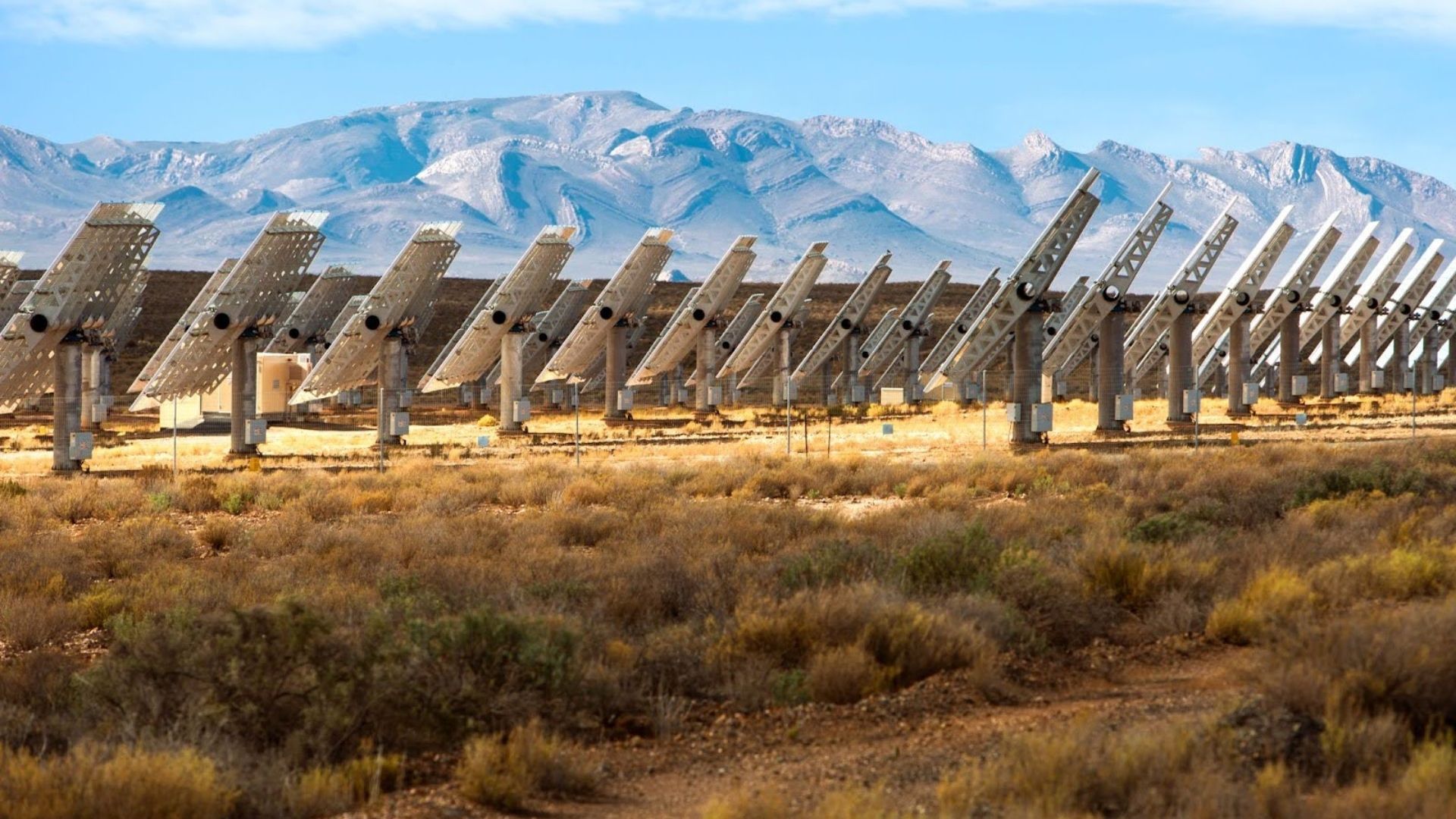 Rows of large solar panels angled toward the sun in a dry, grassy desert landscape with mountain range under blue sky in the background.
