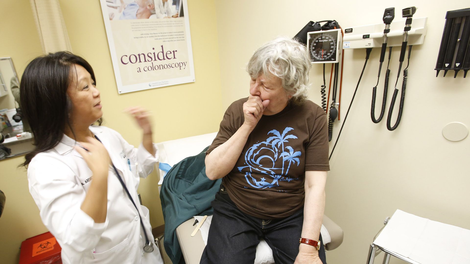 A patient sits on an exam table and coughs while a physician looks at her.